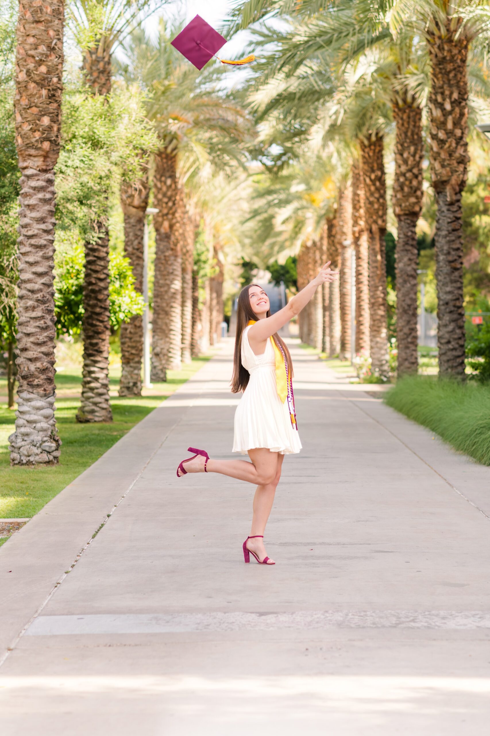 A senior at Arizona State University on Palm Walk throwing her maroon graduation hat into the air with her graduation cords around her neck in a white dress and maroon heals