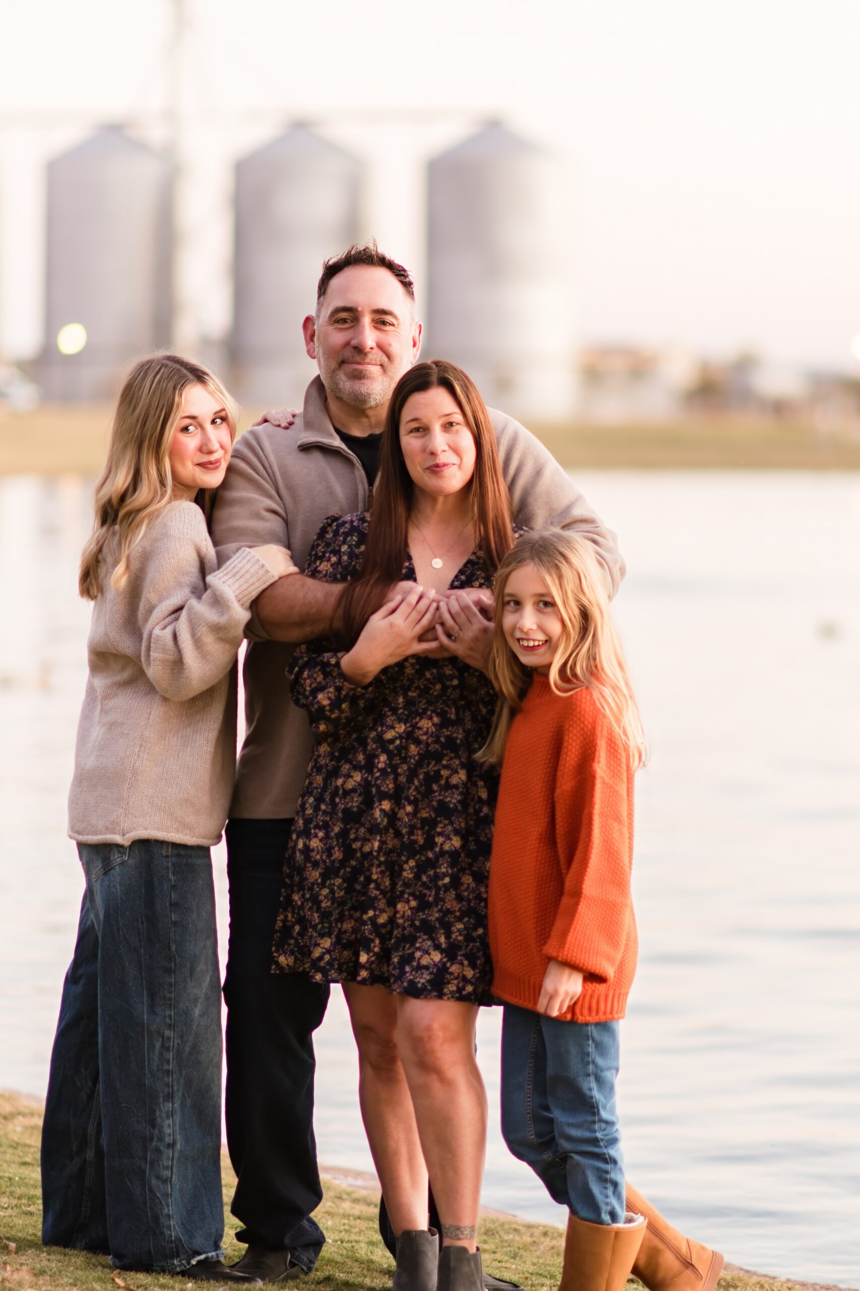 Mom, Dad, and two sisters standing on grass in front of a lake next to the silos in Gilbert, AZ. The girls and father are wearing jeans and a light sweater while the mother is wearing a floral print dress.