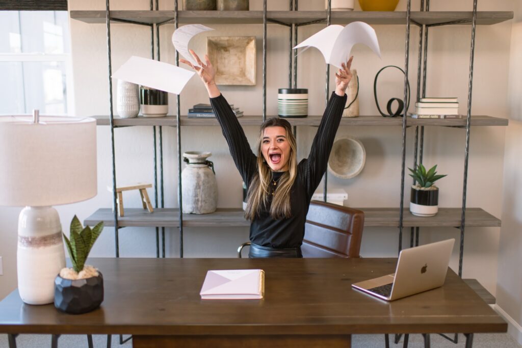 Young female real estate agent sitting at a desk wearing a black outfit throwing papers into the air in celebration.