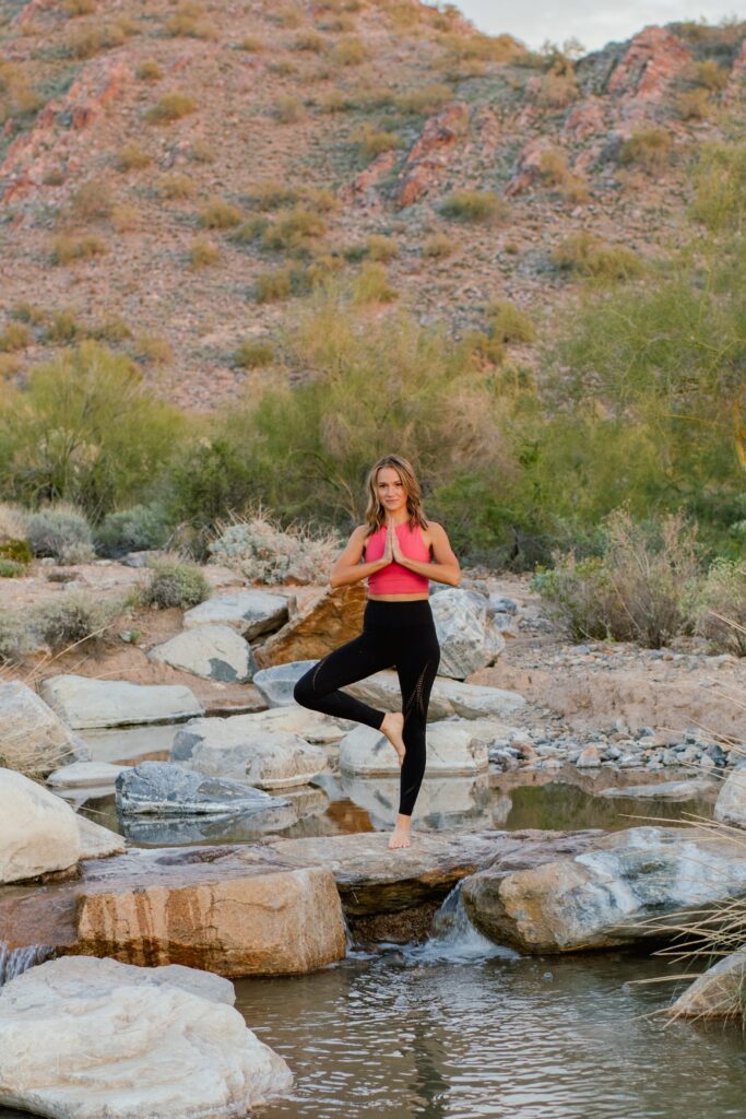 A woman standing on the rocks in the tree pose wearing a pink top and black tights