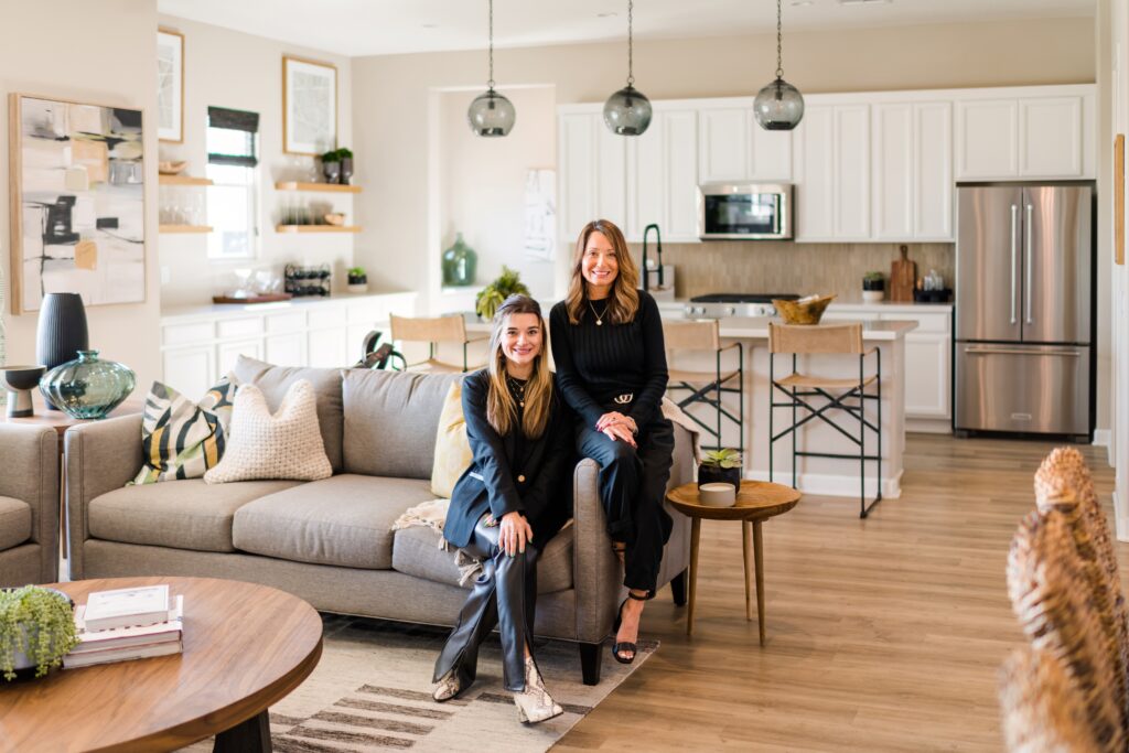 Mother and daughter real estate agents sitting in a home on a couch on cushion one on the arm of the couch smiling at the camera.