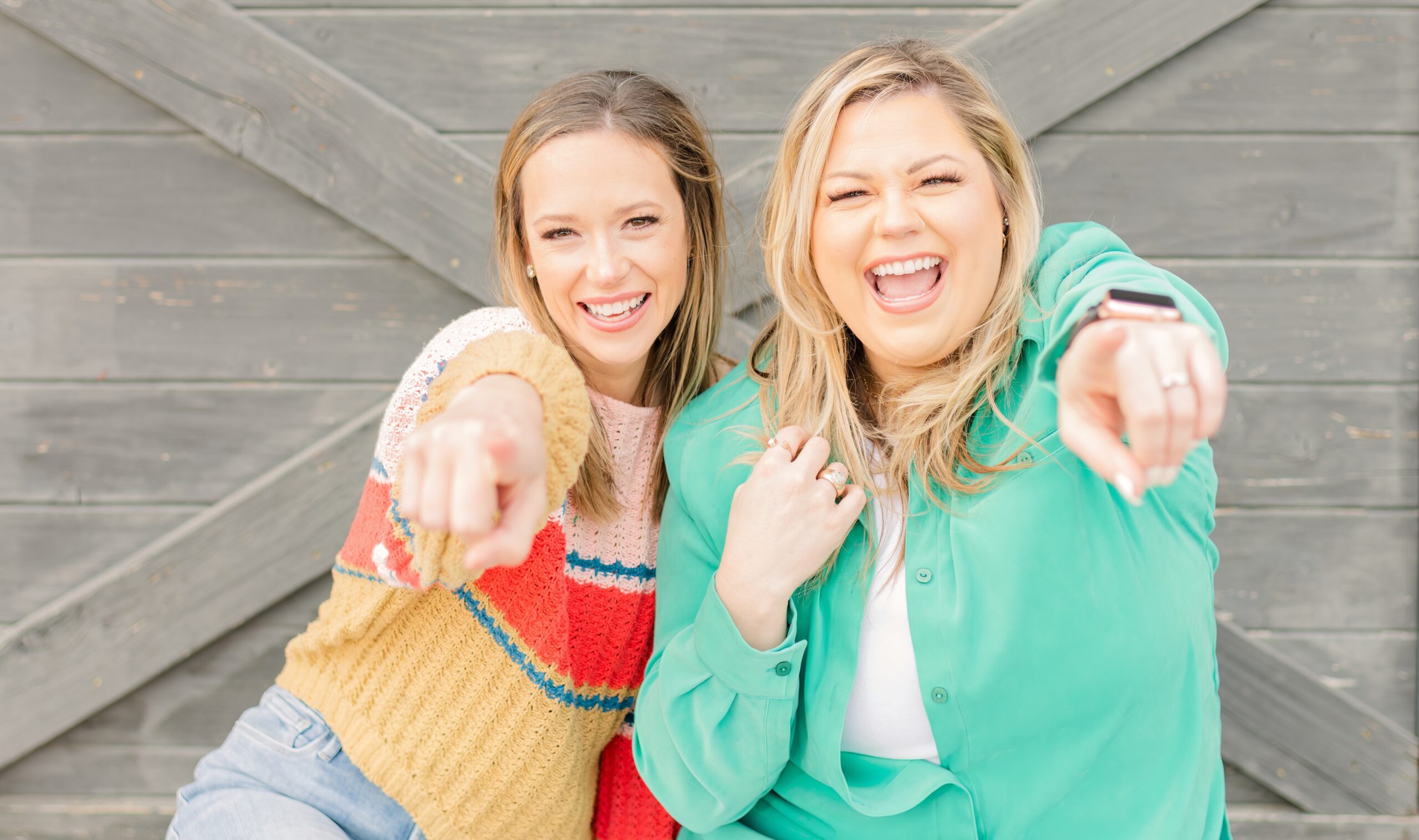 Two female podcasters pointing towards the camera their pointer finger with very big smiles on their face. One is wearing a green shirt and the other is wearing a striped sweater.