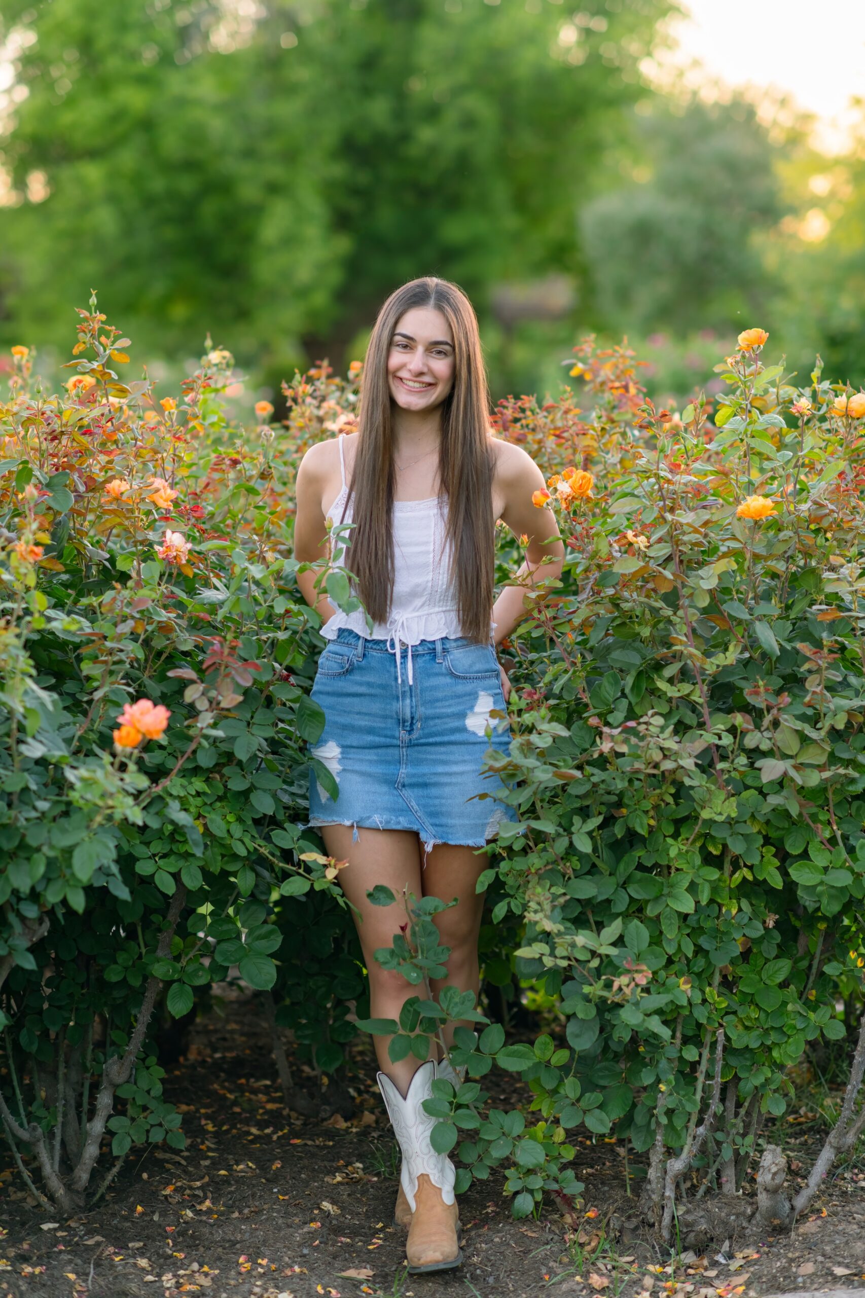 Senior girl at the Mesa Community College Rose Garden, wearing a white top a denim skirt and cowboy boots.