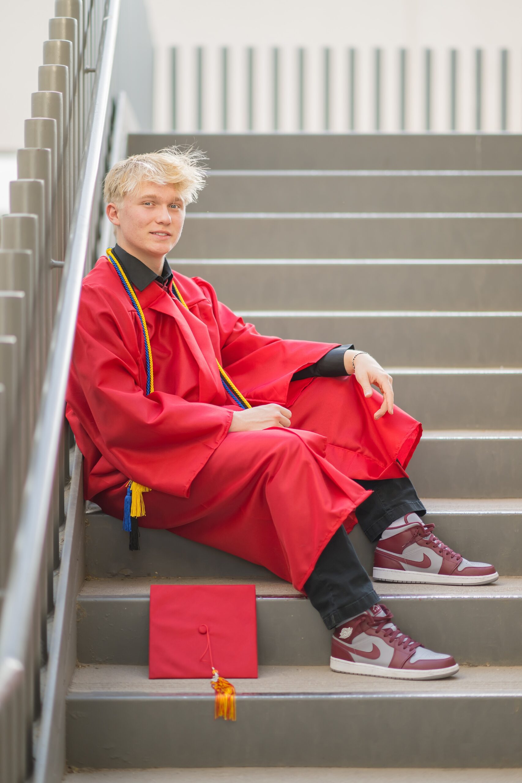 boy sitting on a stair case in gown with cap next to him, one leg up and one leg down