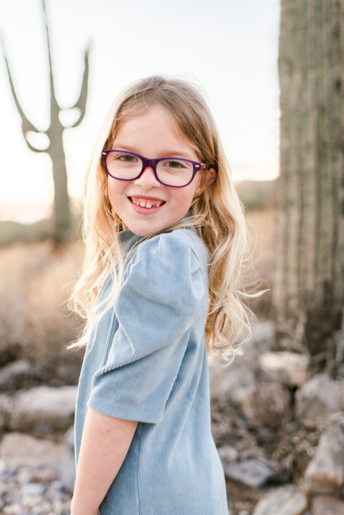 A little girl with a blue sweater dress on and purple glasses smiling at the camera in the arizona desert