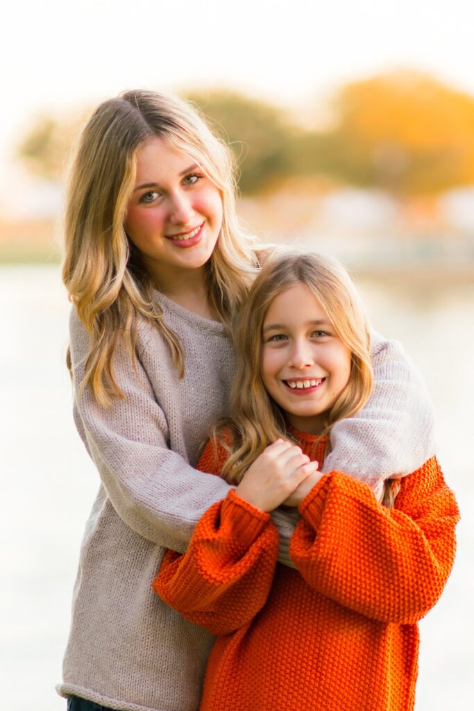 two sisters standing together with the older sister with her hands around the younger sister. The older sister is wearing a tan sweater while the younger sister is wearing an orange sweater.