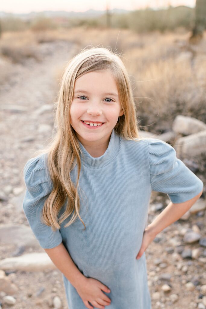 A little girl with a blue sweater dress on smiling at the camera in the arizona desert 