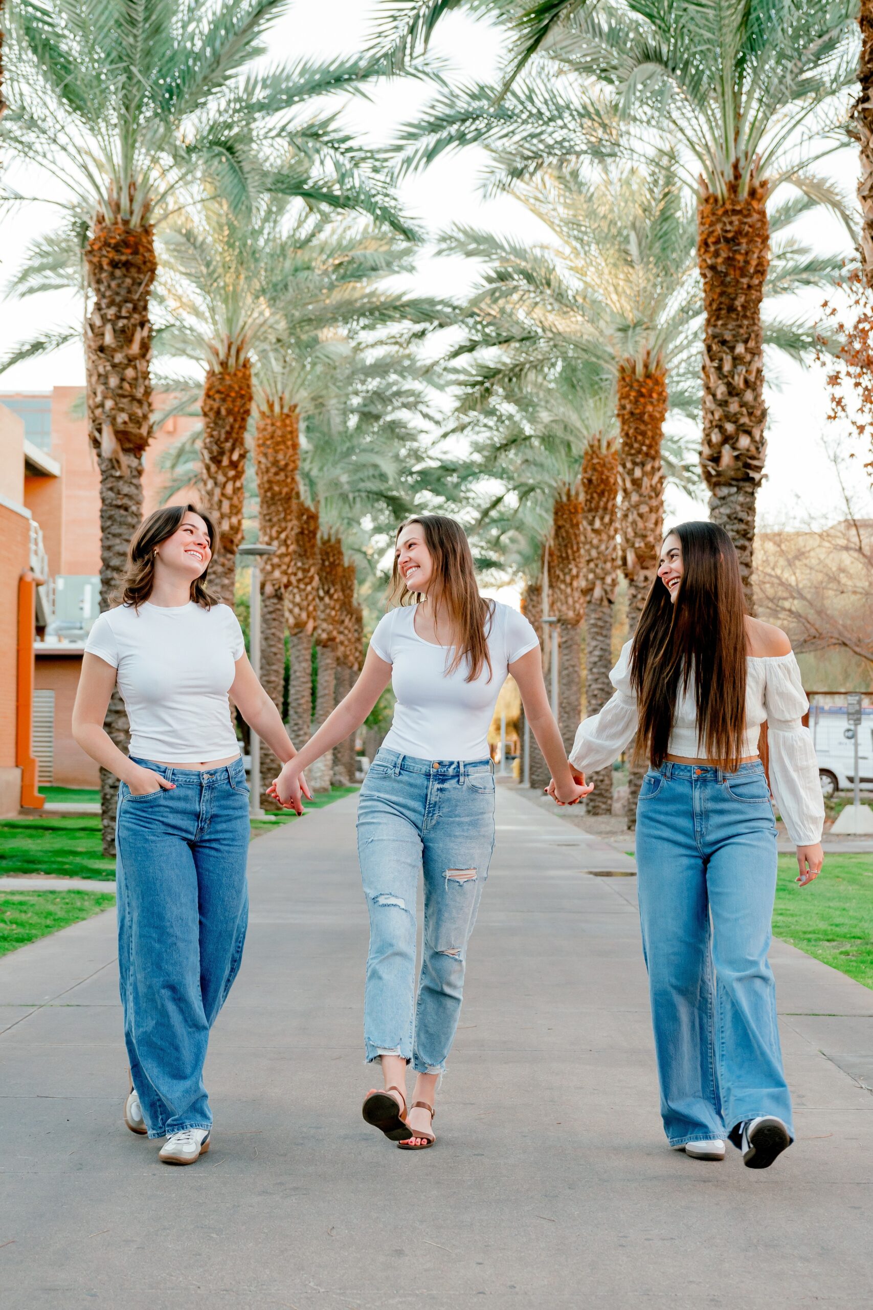 A group of three women hold hands while walking a sidewalk lined with palm trees