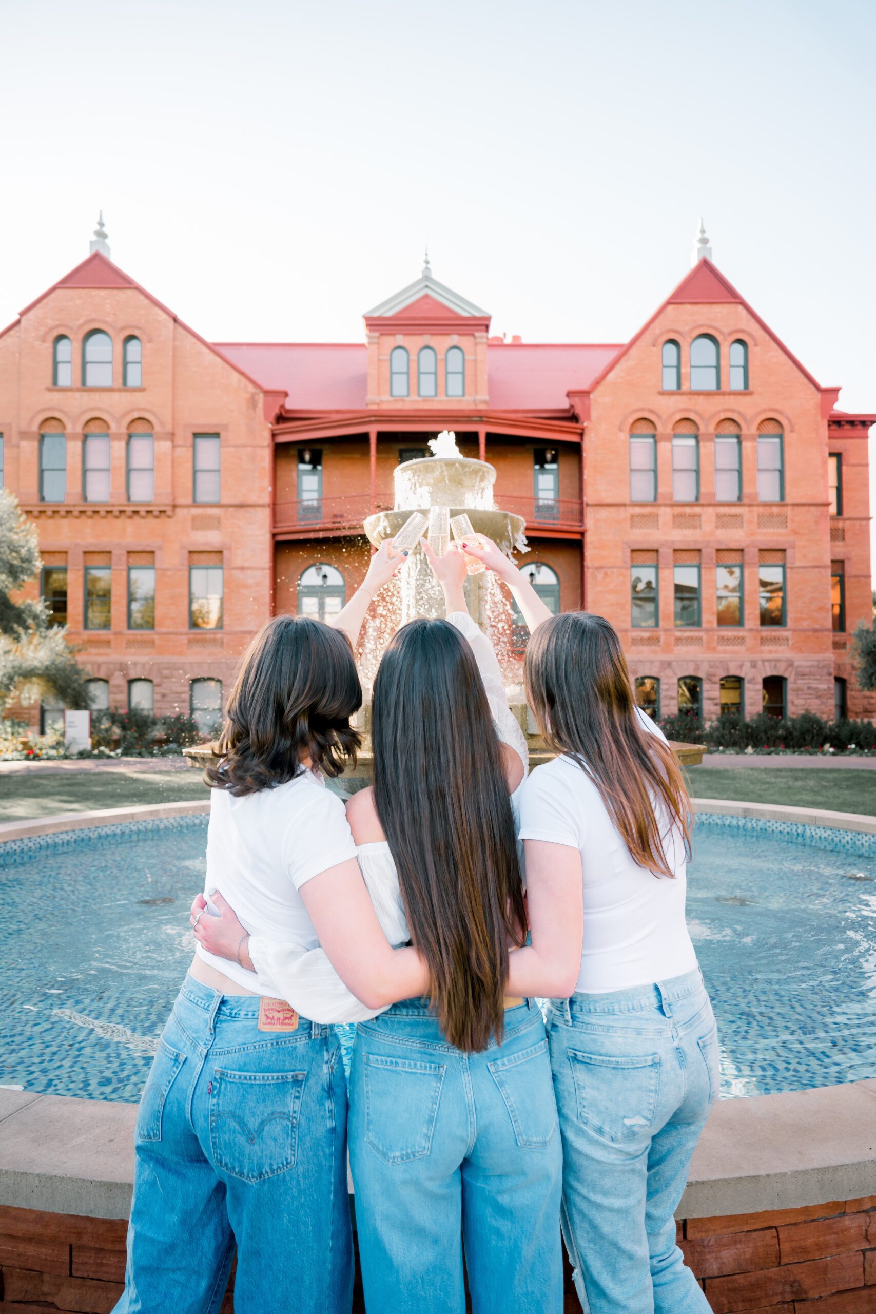 Three girls cheers champagne glasses at the fountain in jeans and white shirts