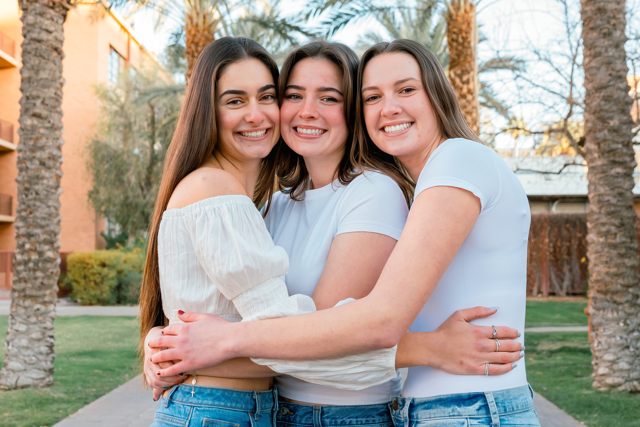 Three girlfriends hug and smile in the quad sidewalk in white tops and jeans for their ASU graduation photos