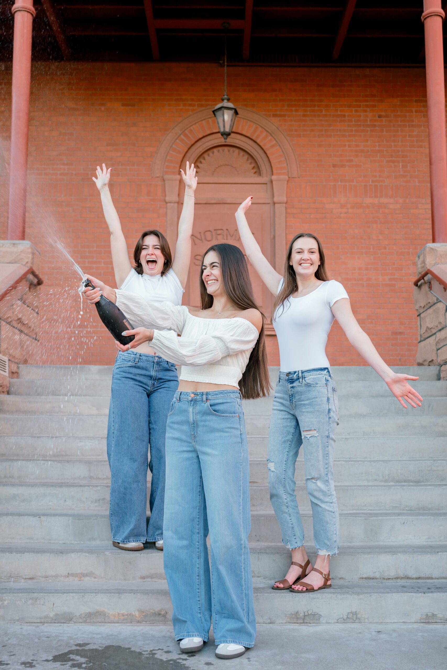 A woman pops champagne as her two friends cheer behind her on steps in white tops and jeans for their ASU graduation photos