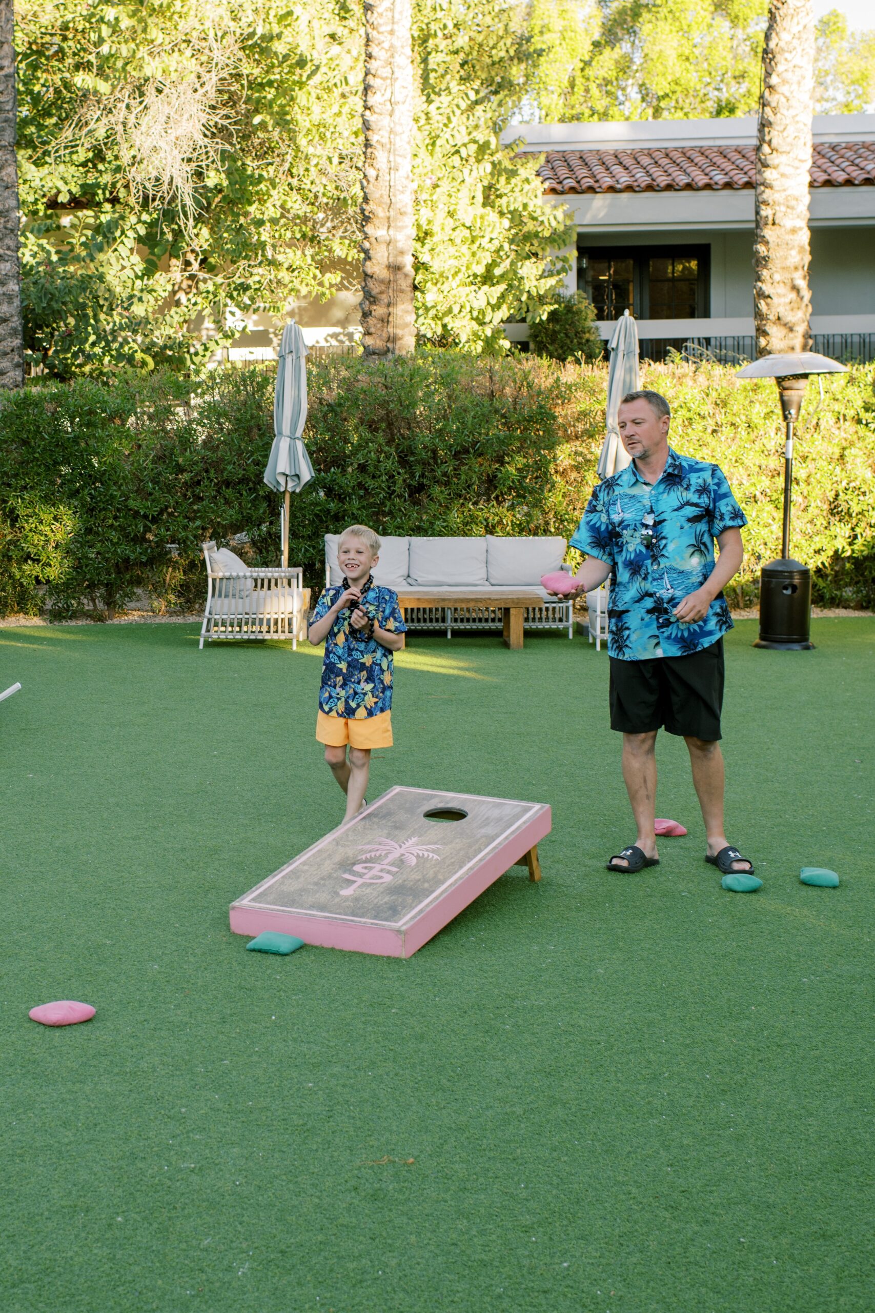 A dad plays corn hole with his young son in matching tropical shirts