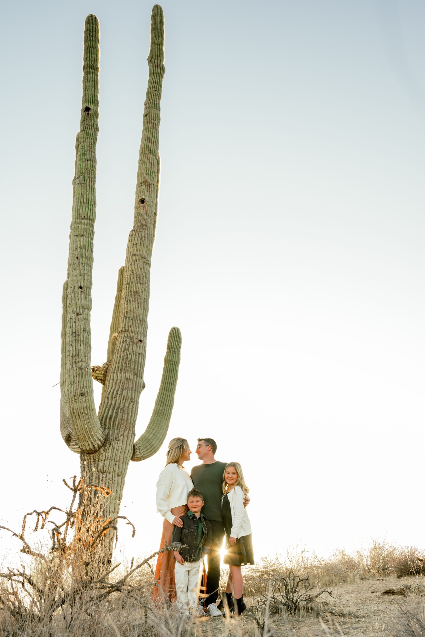 A happy family of 4 smile at each other in a desert trail behind a tall Saguaro cactus