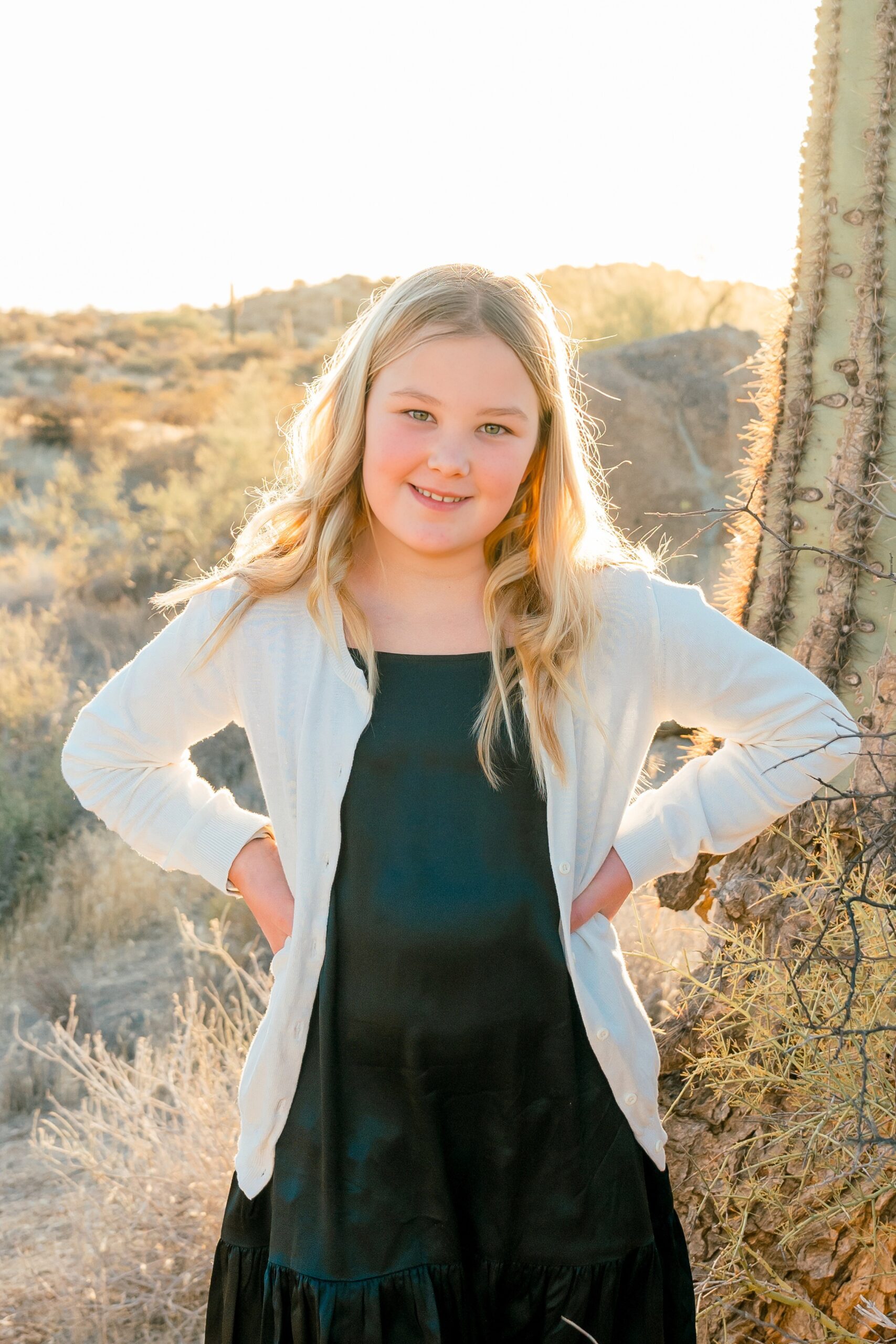 A happy young girl in a green dress and white sweater stands with hands on her hip in a desert trail