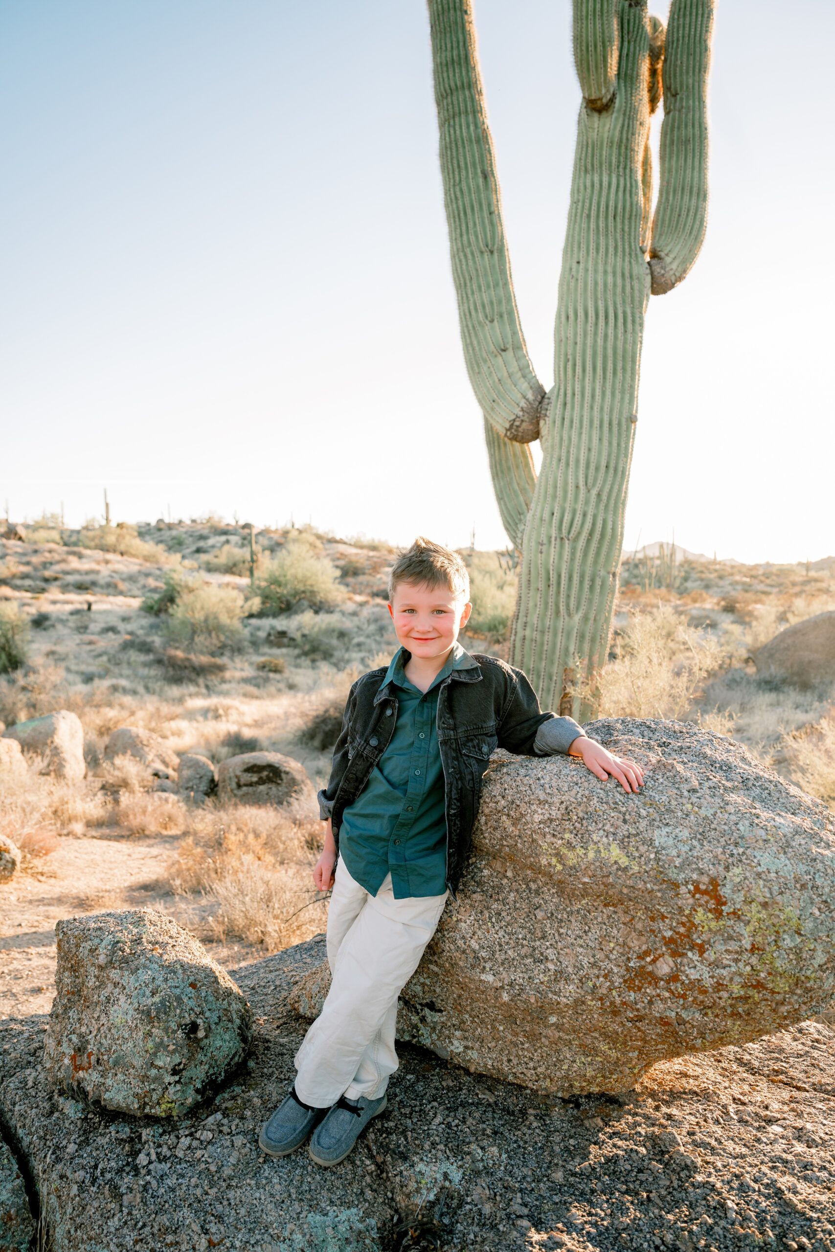 A happy young boy in white pants and denim jacket leans on a boulder under a Saguaro cactus at sunset during family things to do in scottsdale