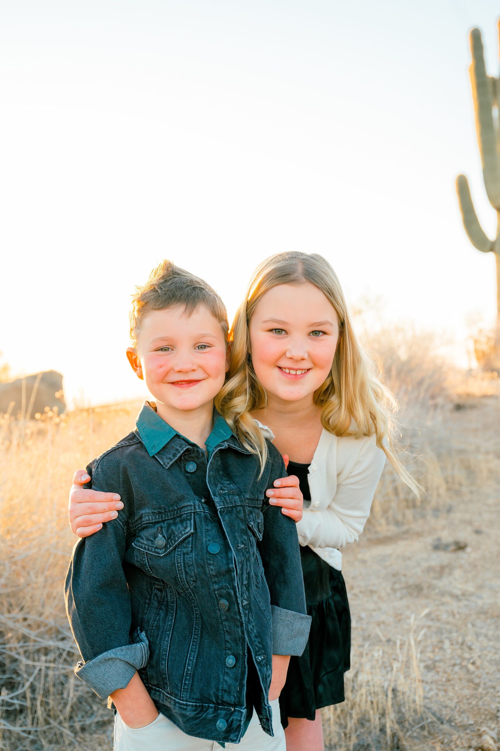 Happy young brother and sister in a denim jacket and white sweater smile in a desert trail at sunset