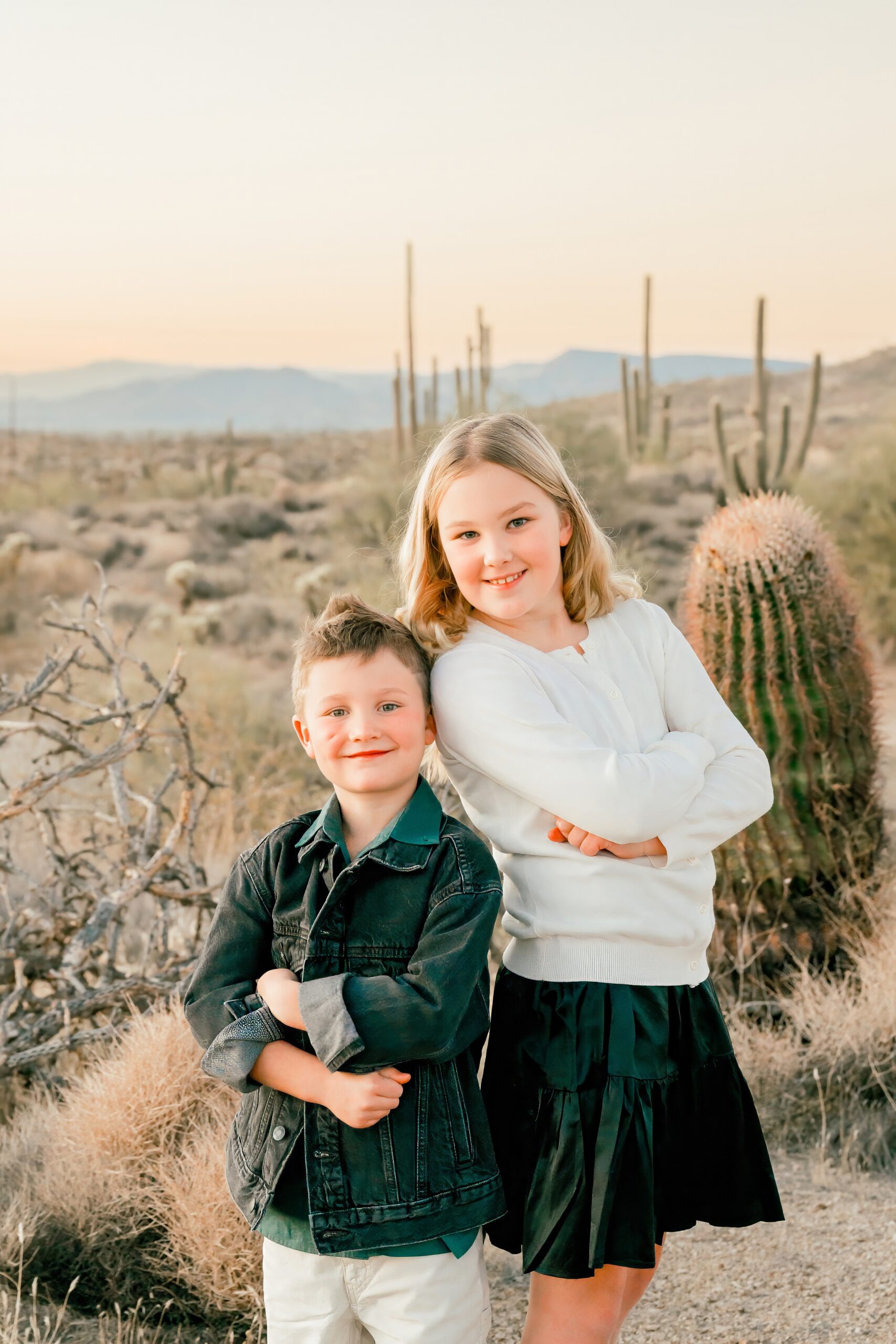 A young girl leans on her little brother both with arms crossed in a desert park at sunset during family things to do in scottsdale