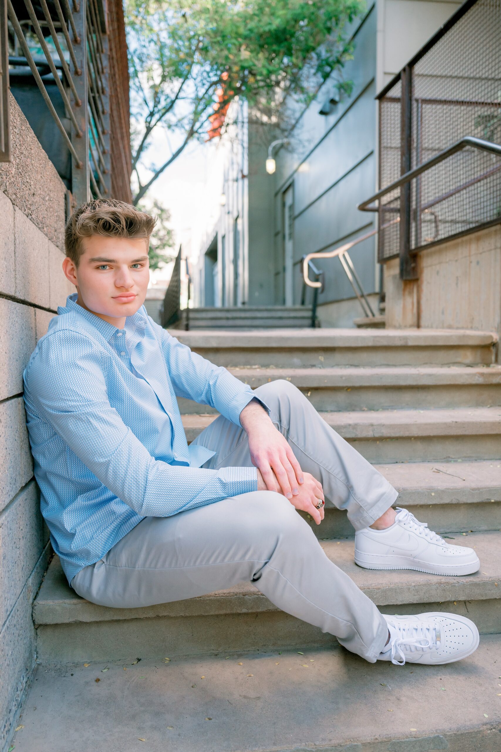 A teenage boy in a blue button down and grey pants sits on concrete steps leaning on his thighs