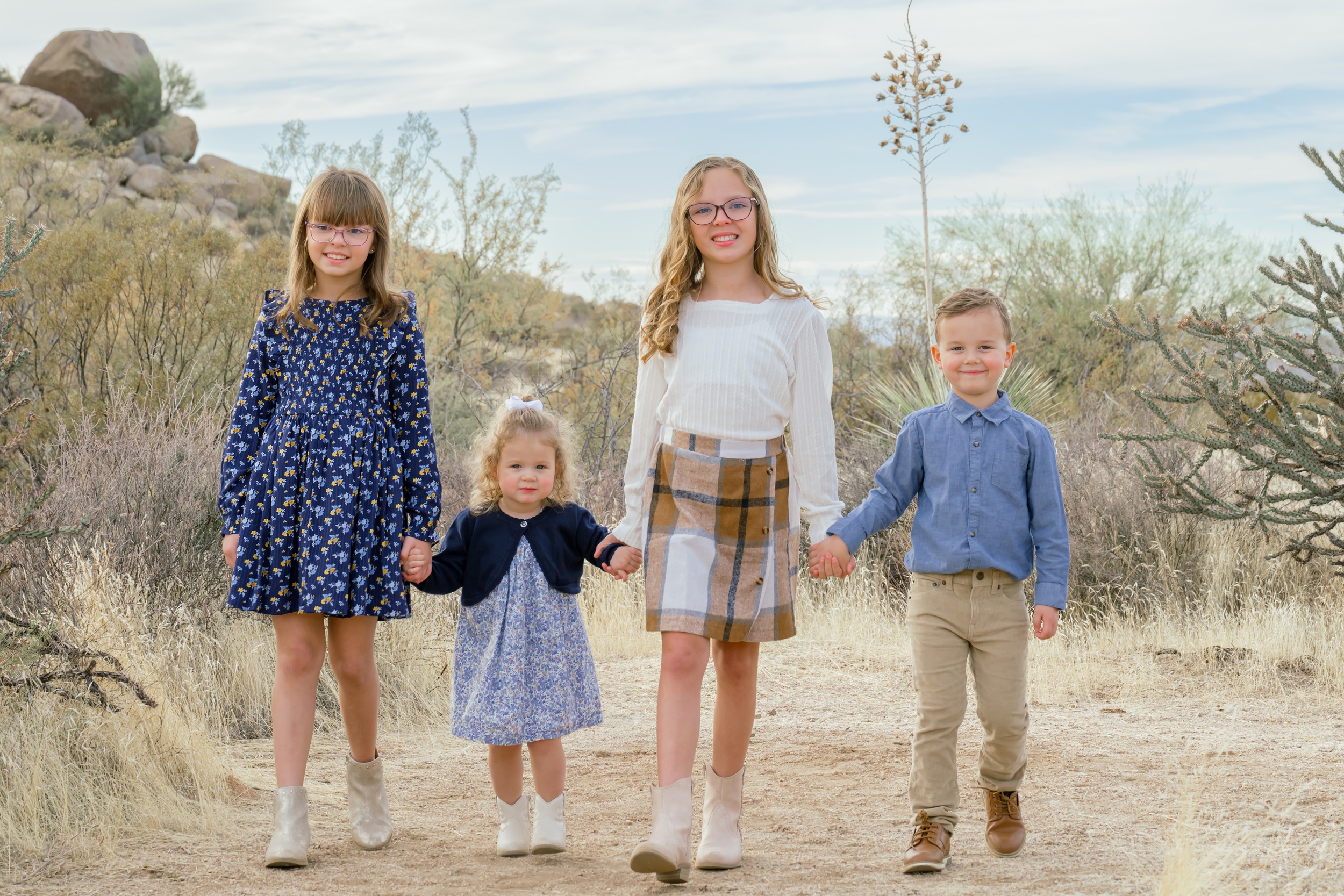 Four young siblings hold hands while walking a desert trail and smiling during scottsdale kid activities