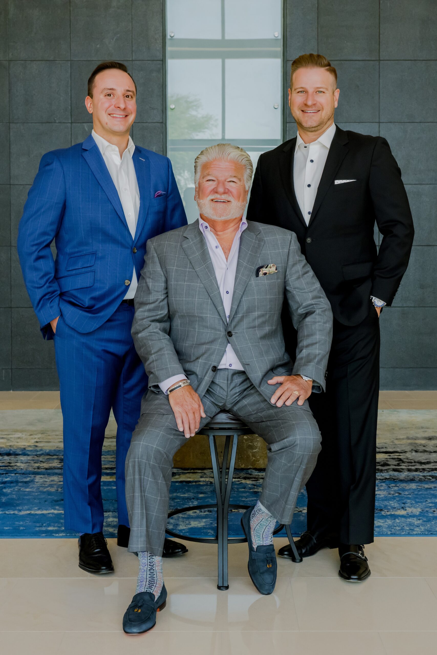Three men in suits stand and sit together in their offices smiling
