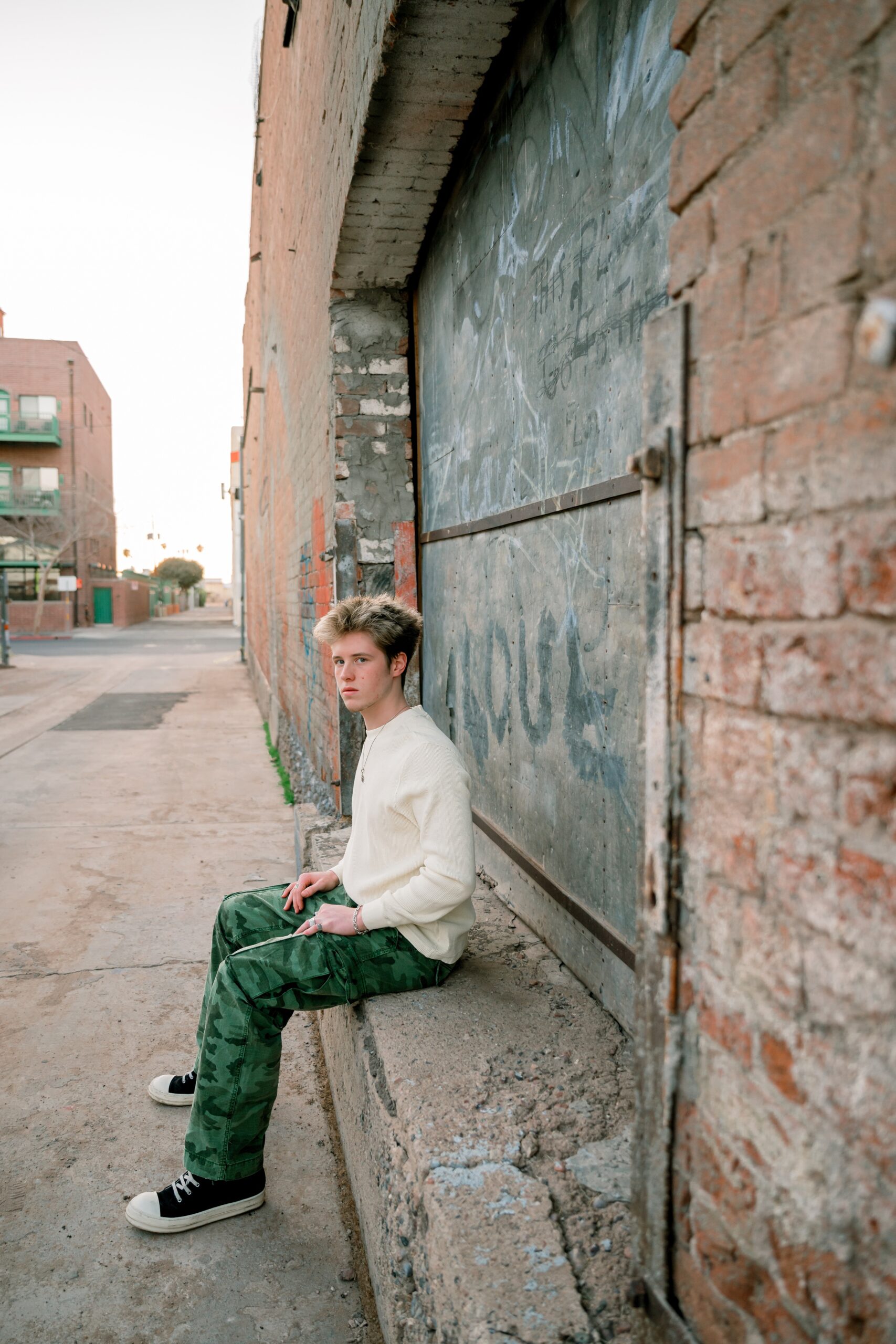 A teenager in camo pants and white shirt sits looking over his shoulder down the sidewalk