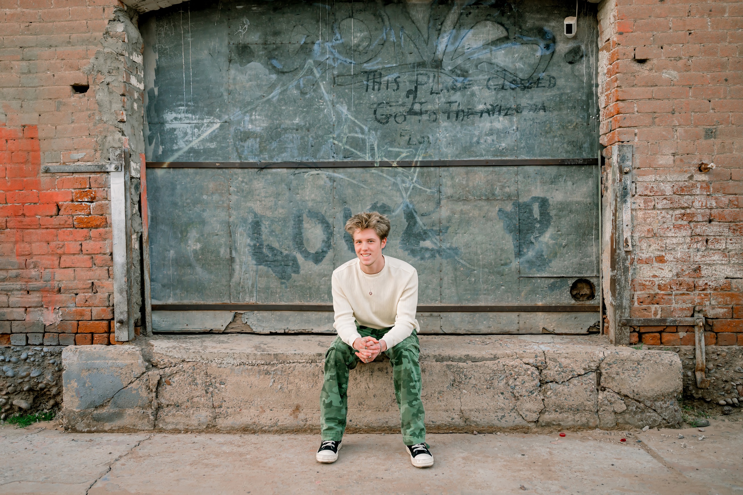A high school senior in a white shirt and camo pants sits on a rustic loading dock