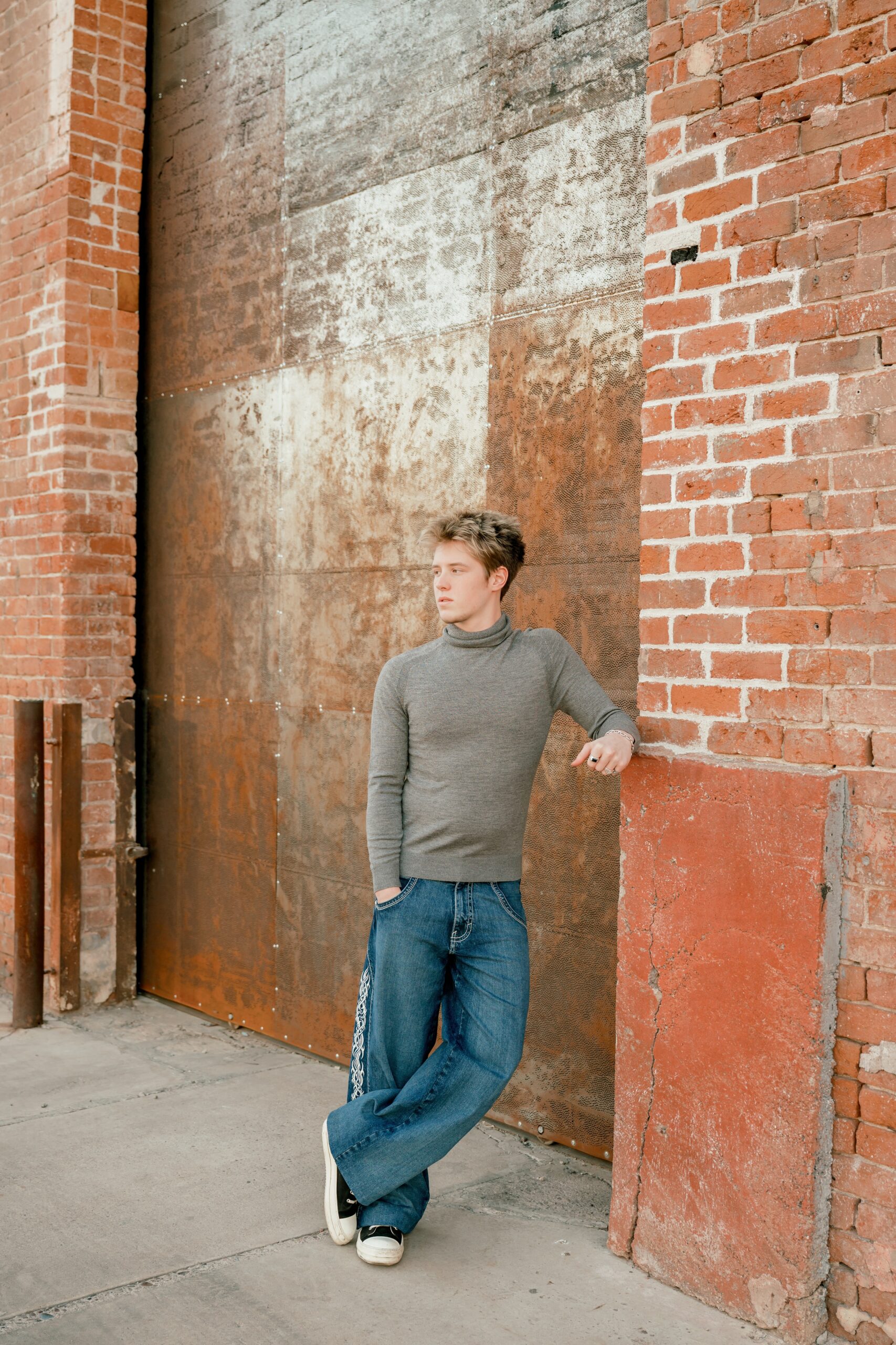A teenager in jeans and brown shirt leans on a brick wall in an alley for some urban senior photos