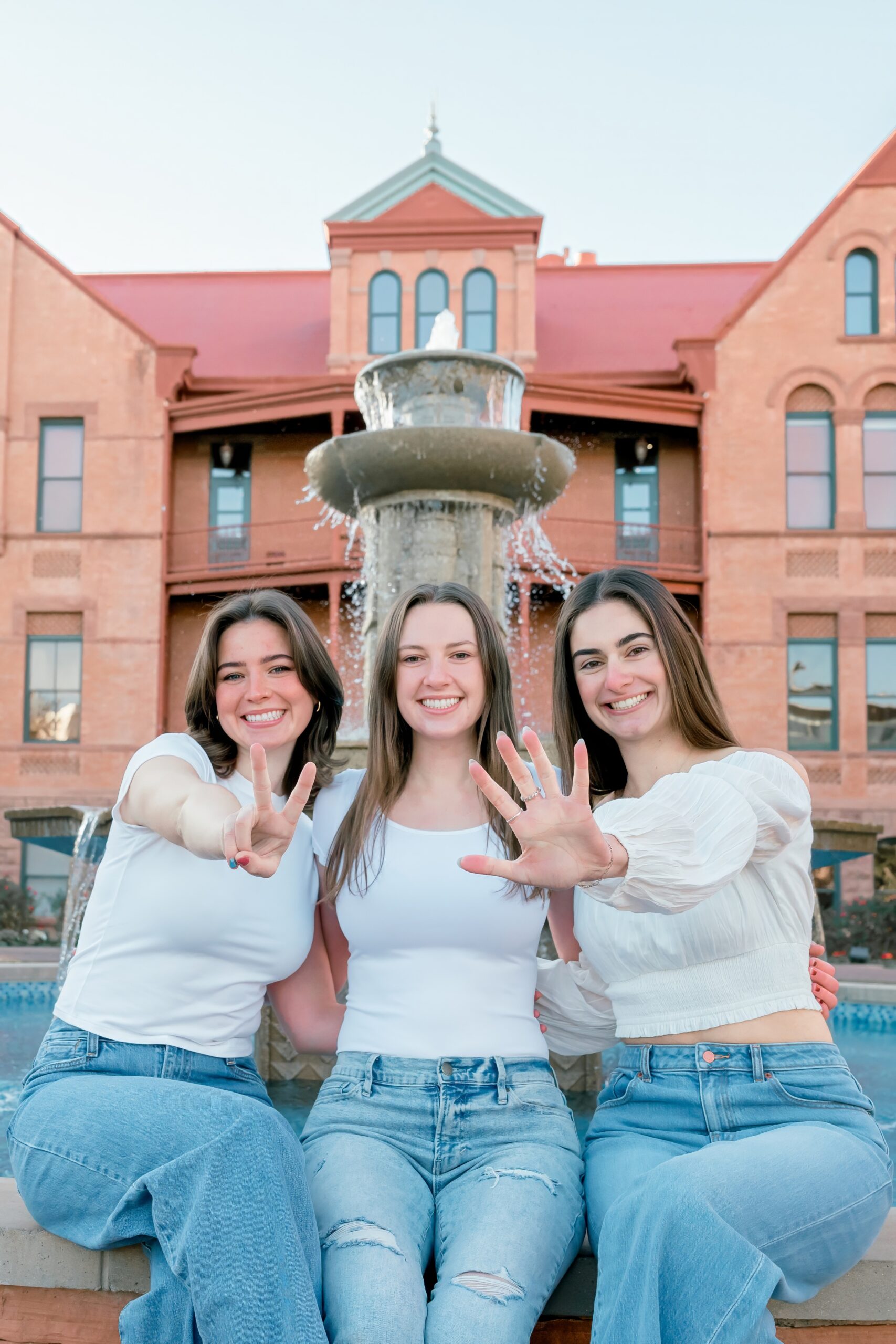 Three women sit on a fountain in matching white shirts and jeans smiling holding out a 25