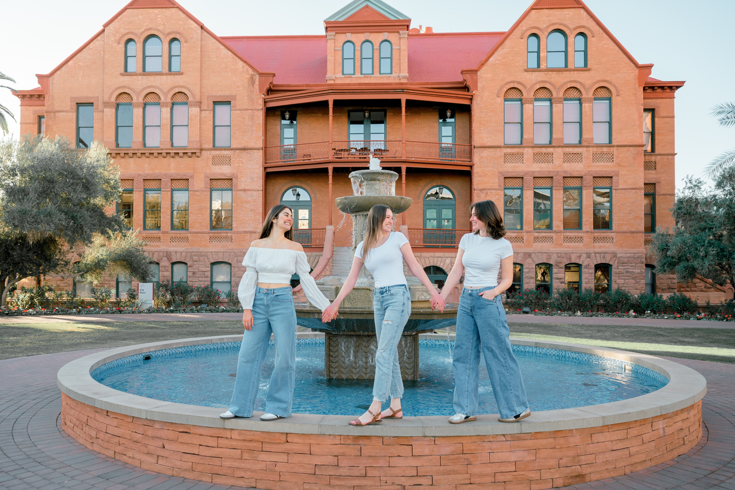Three smiling women walk on a fountain holding hands in jeans and white shirts