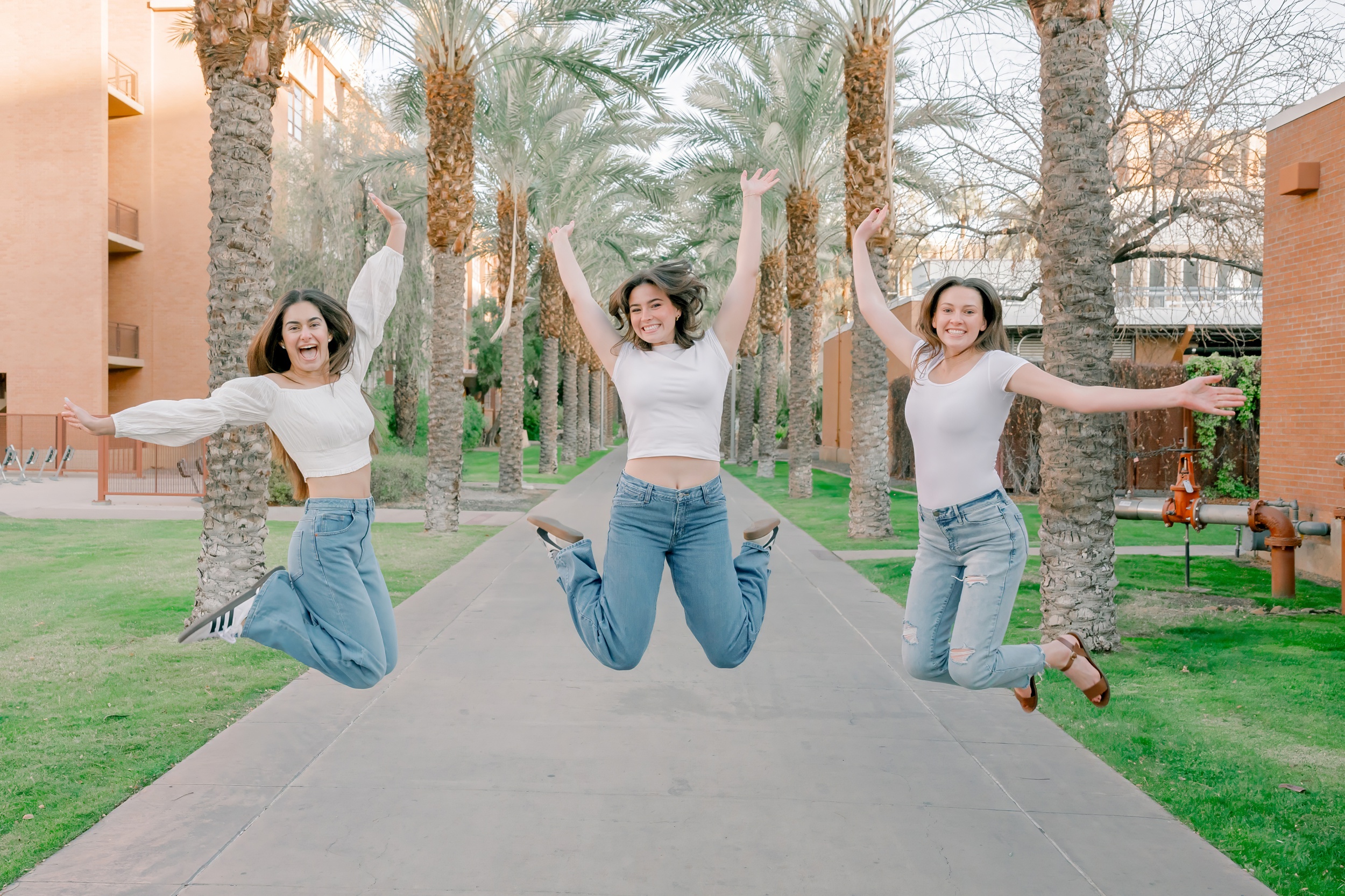 Three happy graduates in white shirts and jeans jump for joy in a palm lined sidewalk after visiting colleges in arizona