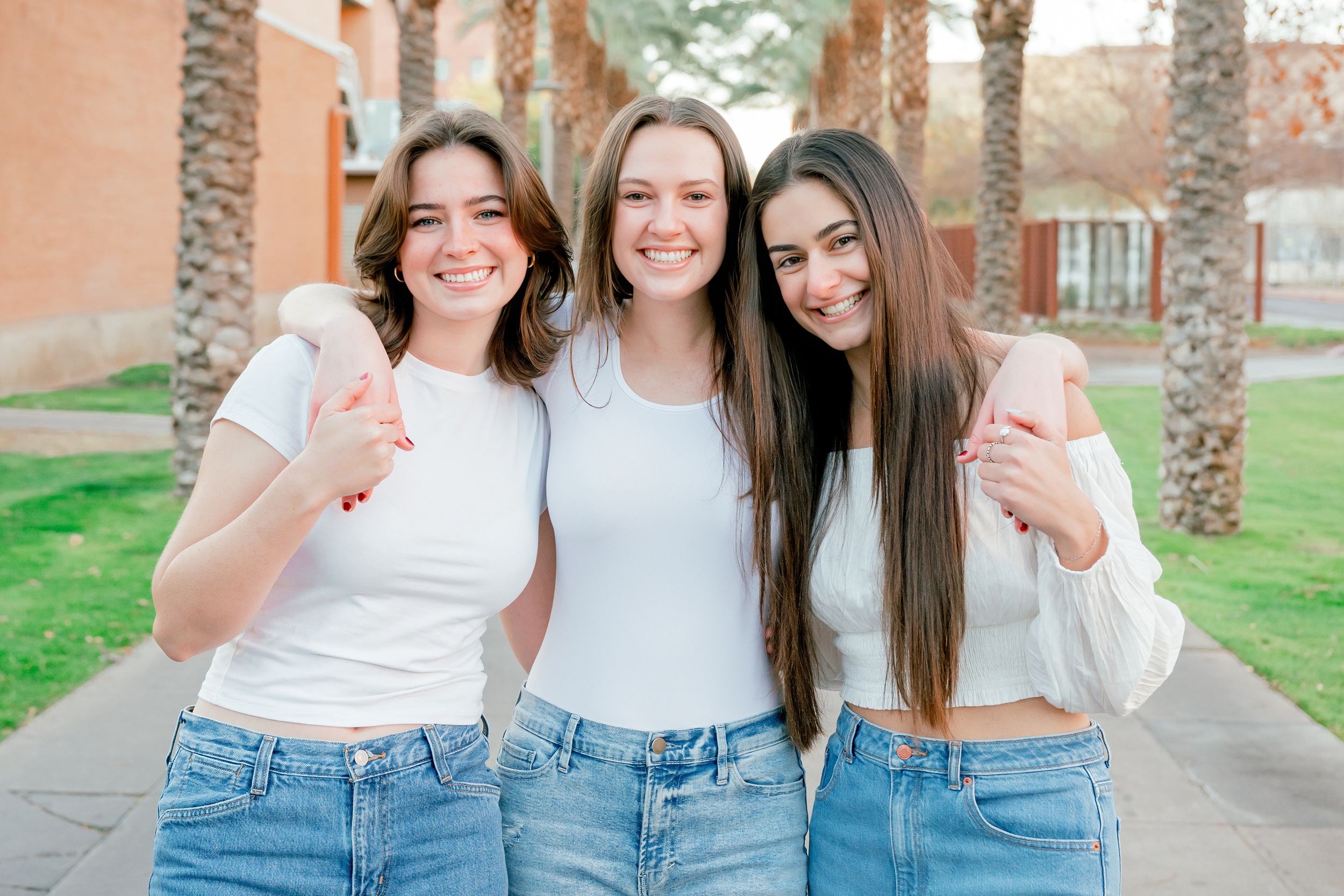 Three happy women in white shirts and jeans smile with arms around each other in a sidewalk after visiting colleges in arizona