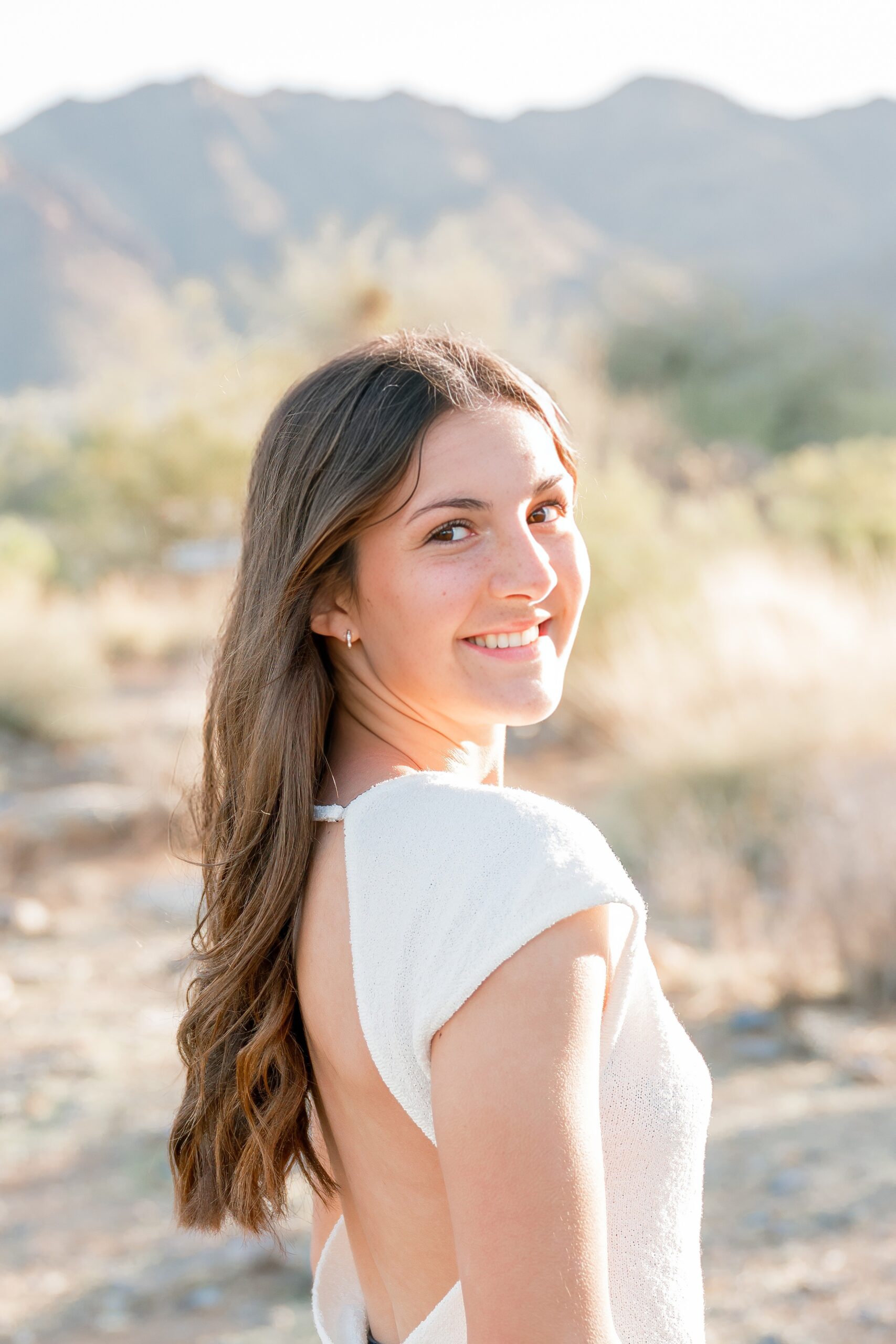 a high school senior in a white dress smiles over her shoulder at sunset after getting hair and makeup in scottsdale, az