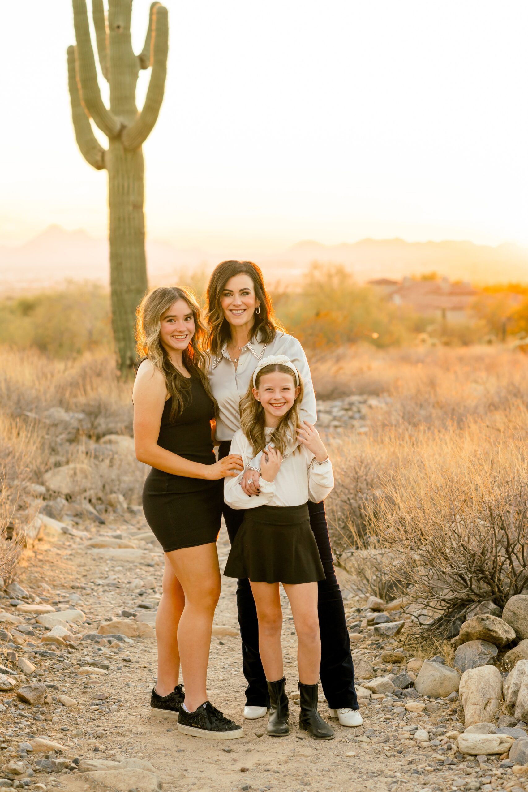 A mom hugs her smiling two daughters in a desert trail at sunset