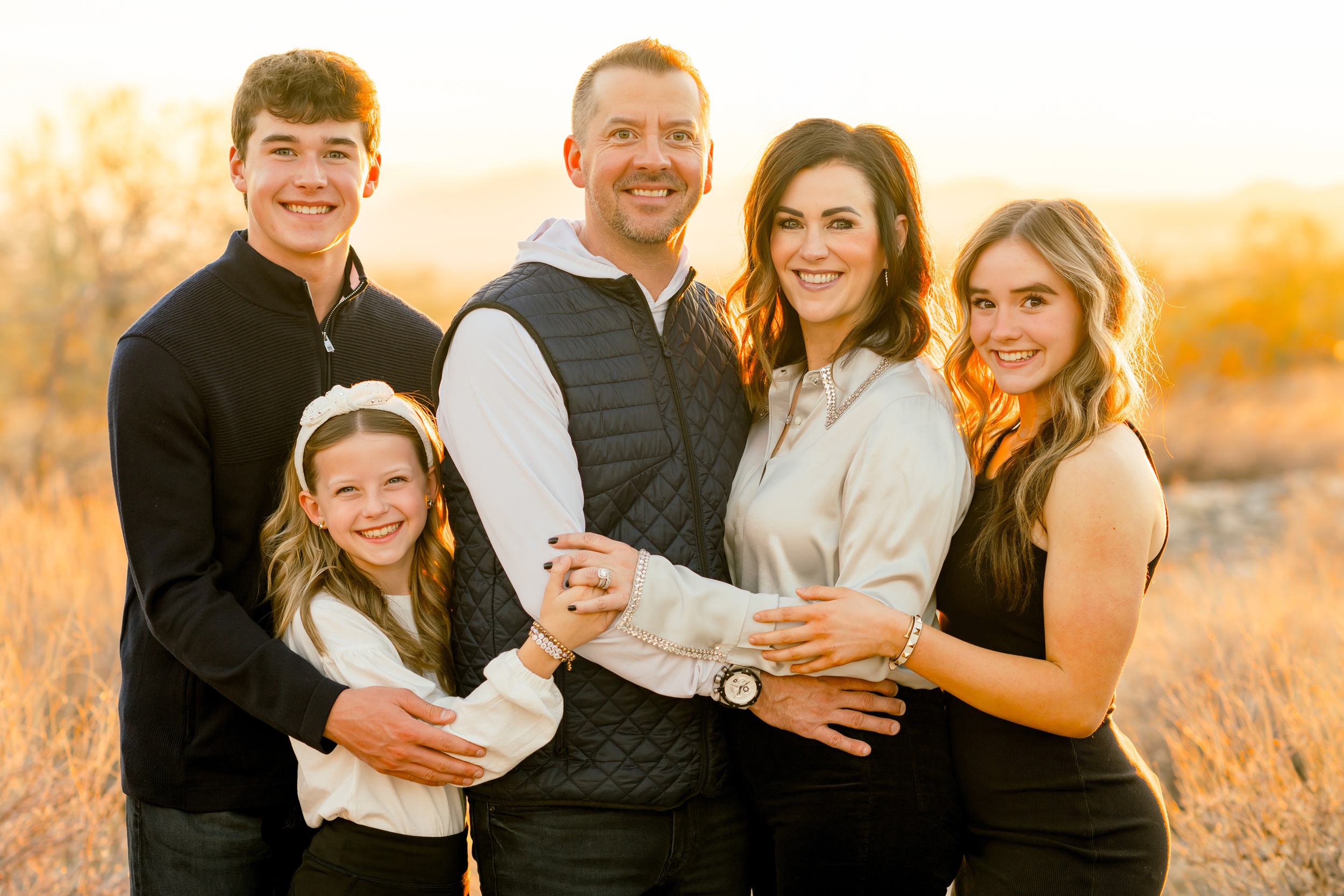 A smiling family of 5 embrace and smile in black and white outfits at sunset