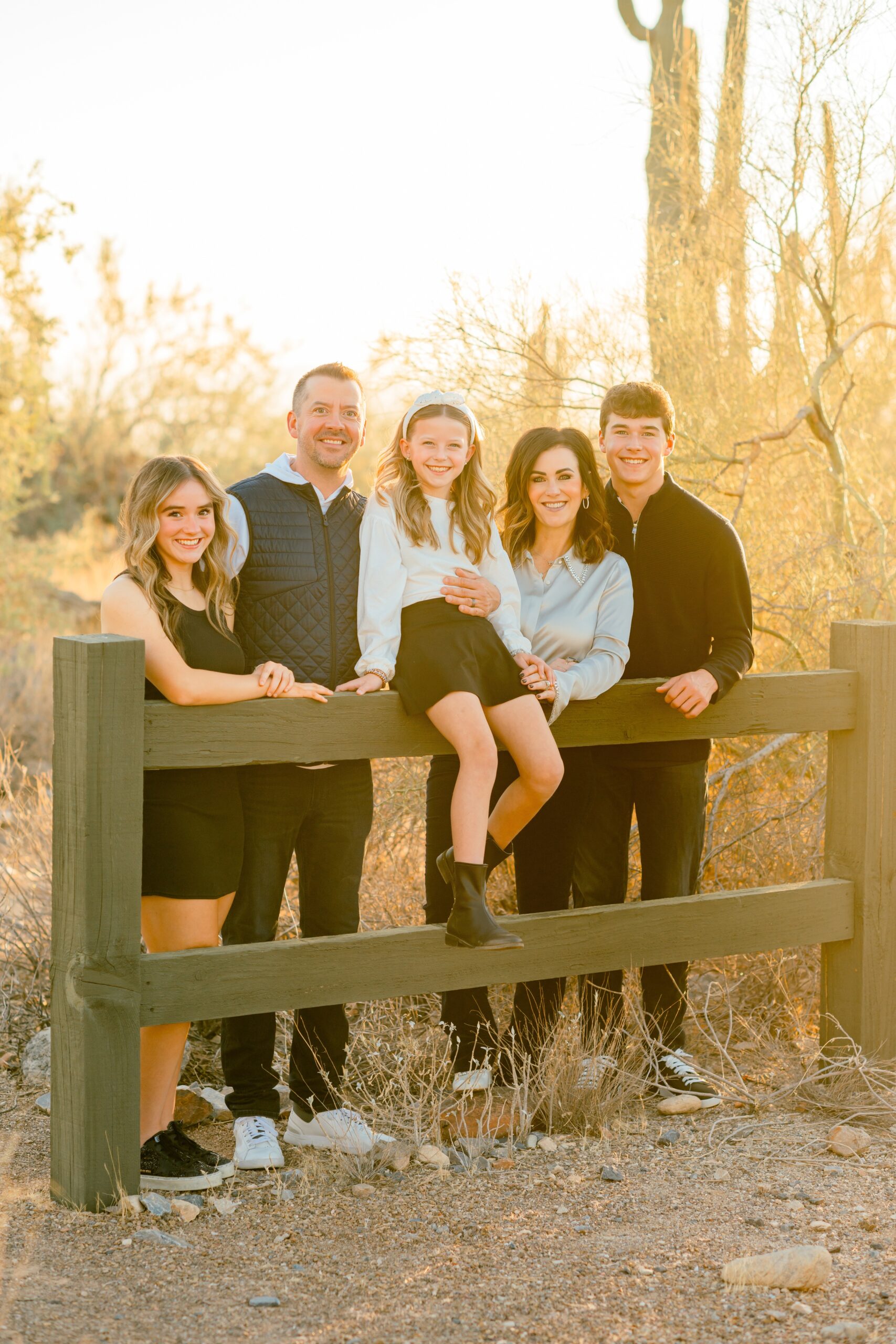 A happy family of 5 lean and sit on a wooden park fence at sunset smiling before visiting indoor playgrounds in scottsdale