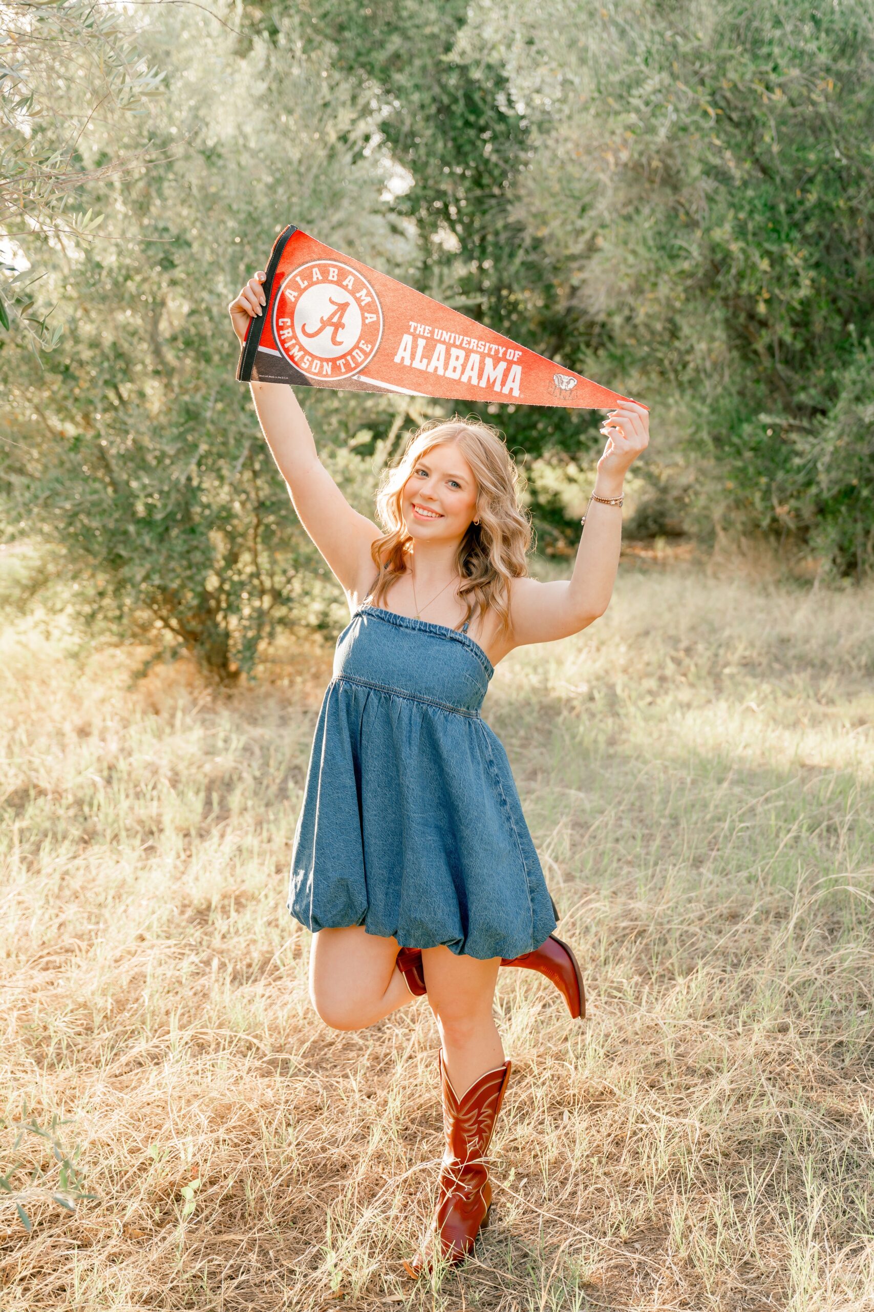 A Desert Mountain High School senior in a denim dress and boots celebrates with a college pennant above her head in a park at sunset