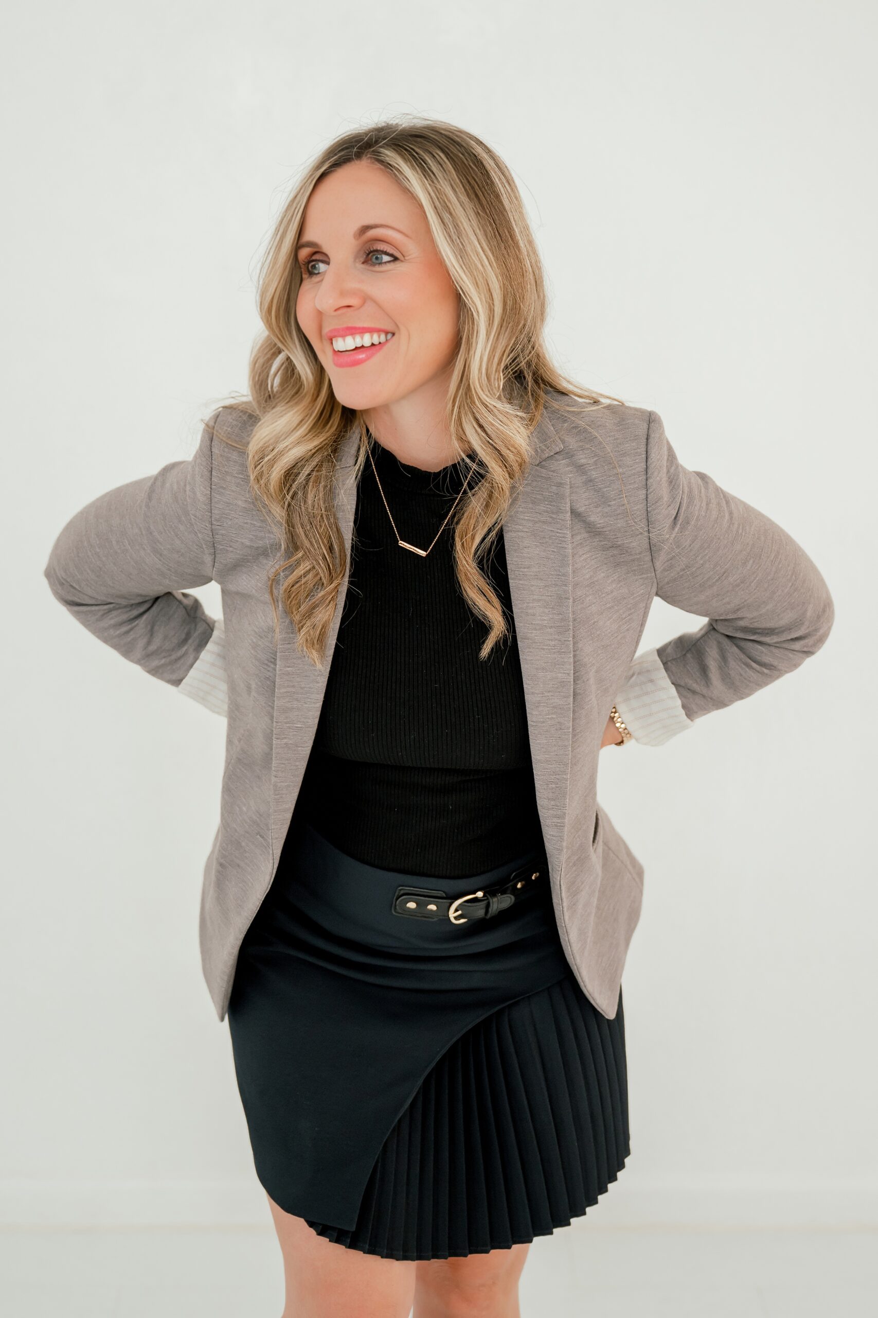 A woman laughs while standing in a studio in a black dress and tan coat