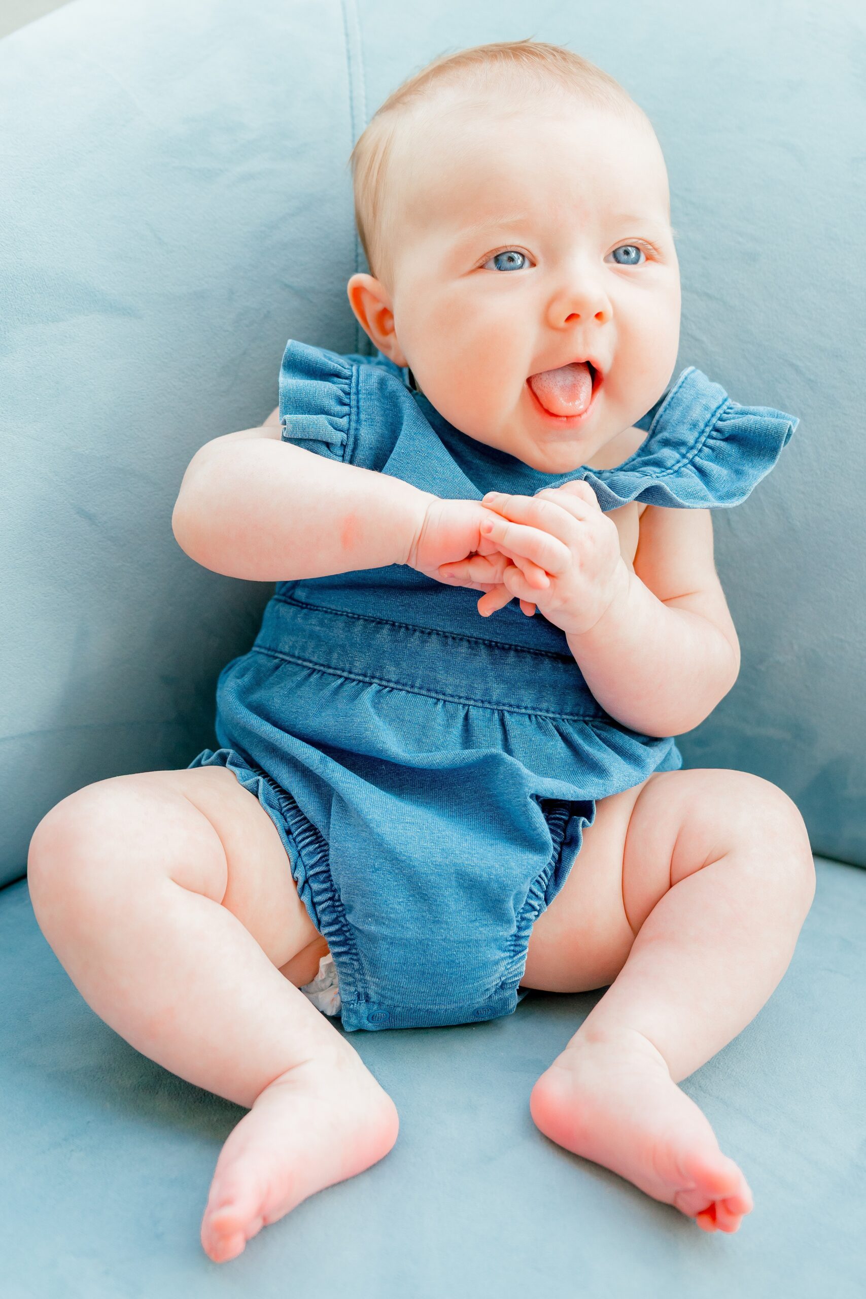 A happy baby in a denim onesie smiles while sitting on a blue couch