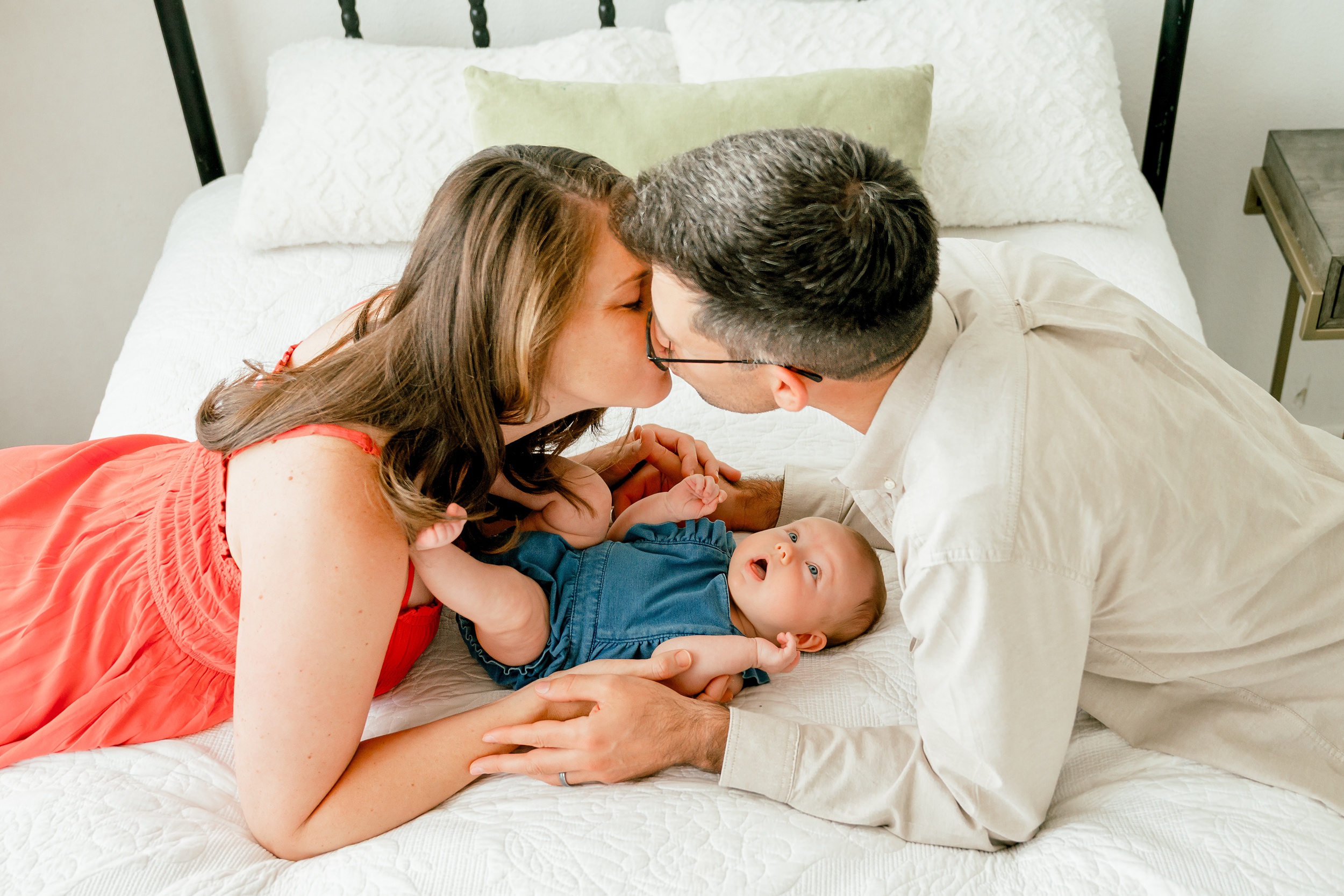 A happy couple kisses over top their laying baby on a bed in a denim onesie in a photography studio in scottsdale az