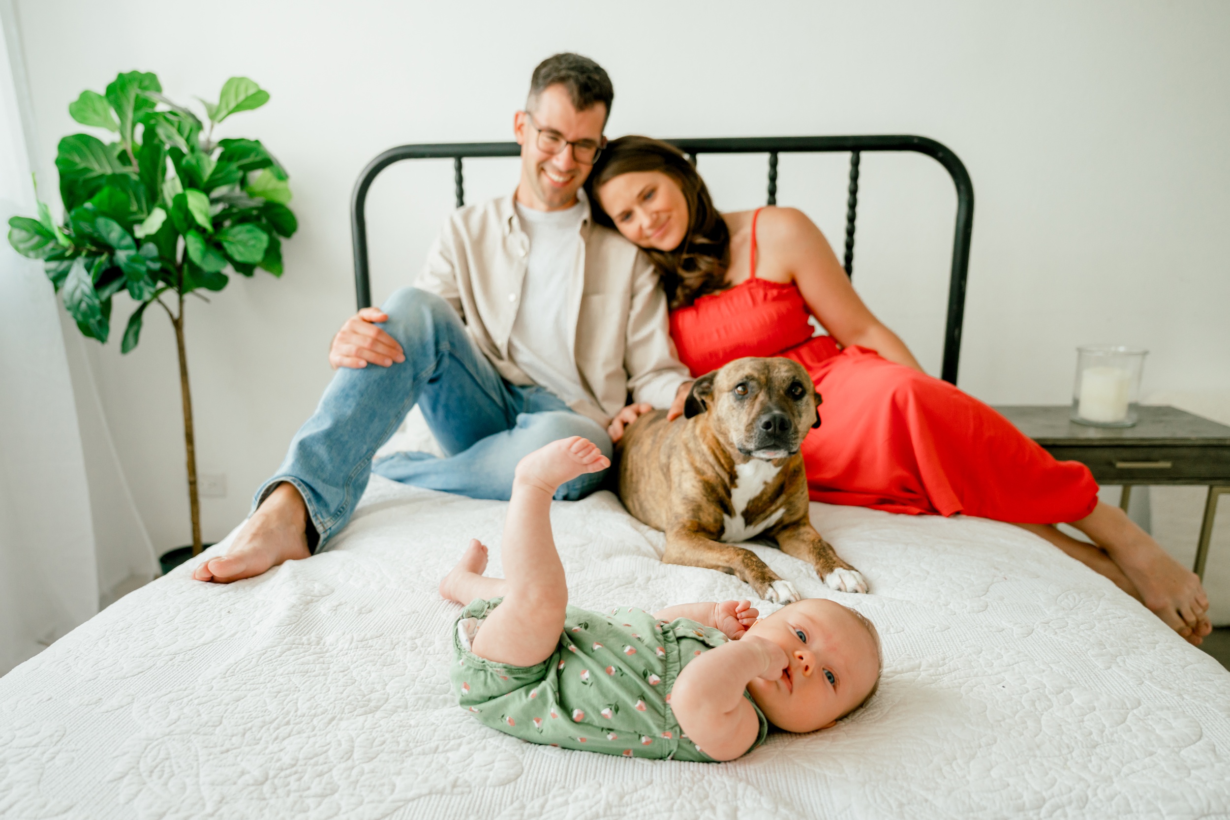 A happy couple and their dog sit on a bed watching over their newborn in a green onesie in a photography studio in scottsdale az