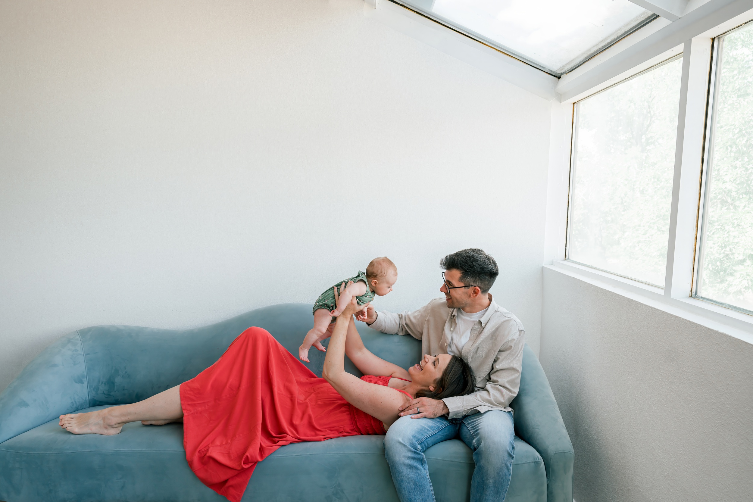 A mom lays resting her head in dad's lap on a couch in a pink dress while they play with their infant above her in a photography studio in scottsdale az