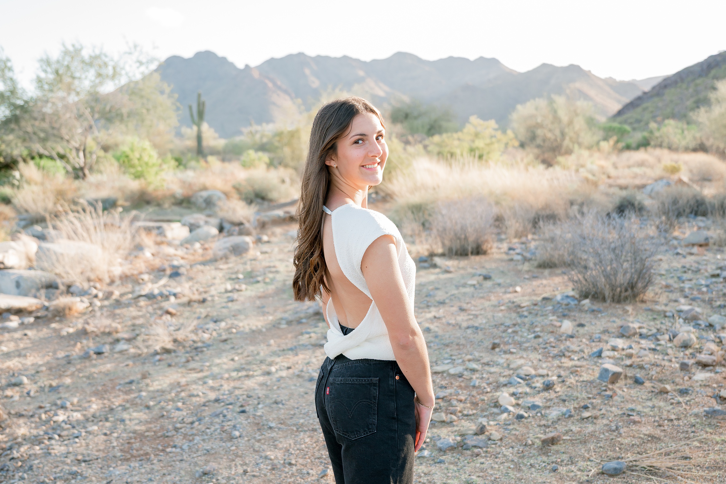 A woman in a white blouse stands smiling over her shoulder in the desert trail