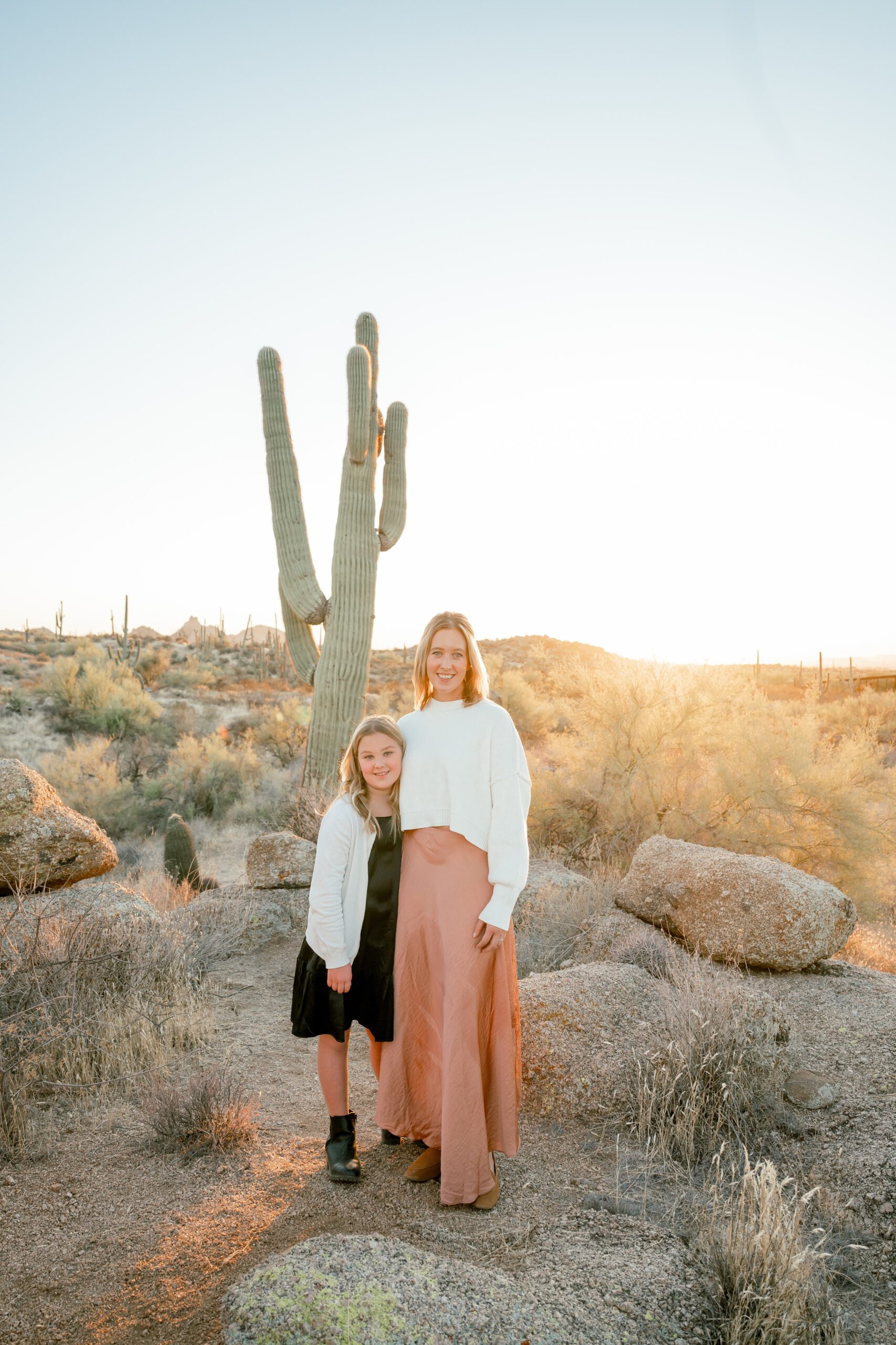 A mom and daughter stand in a desert in white sweaters and pink and black dresses at sunset after visiting womens boutiques in scottsdale