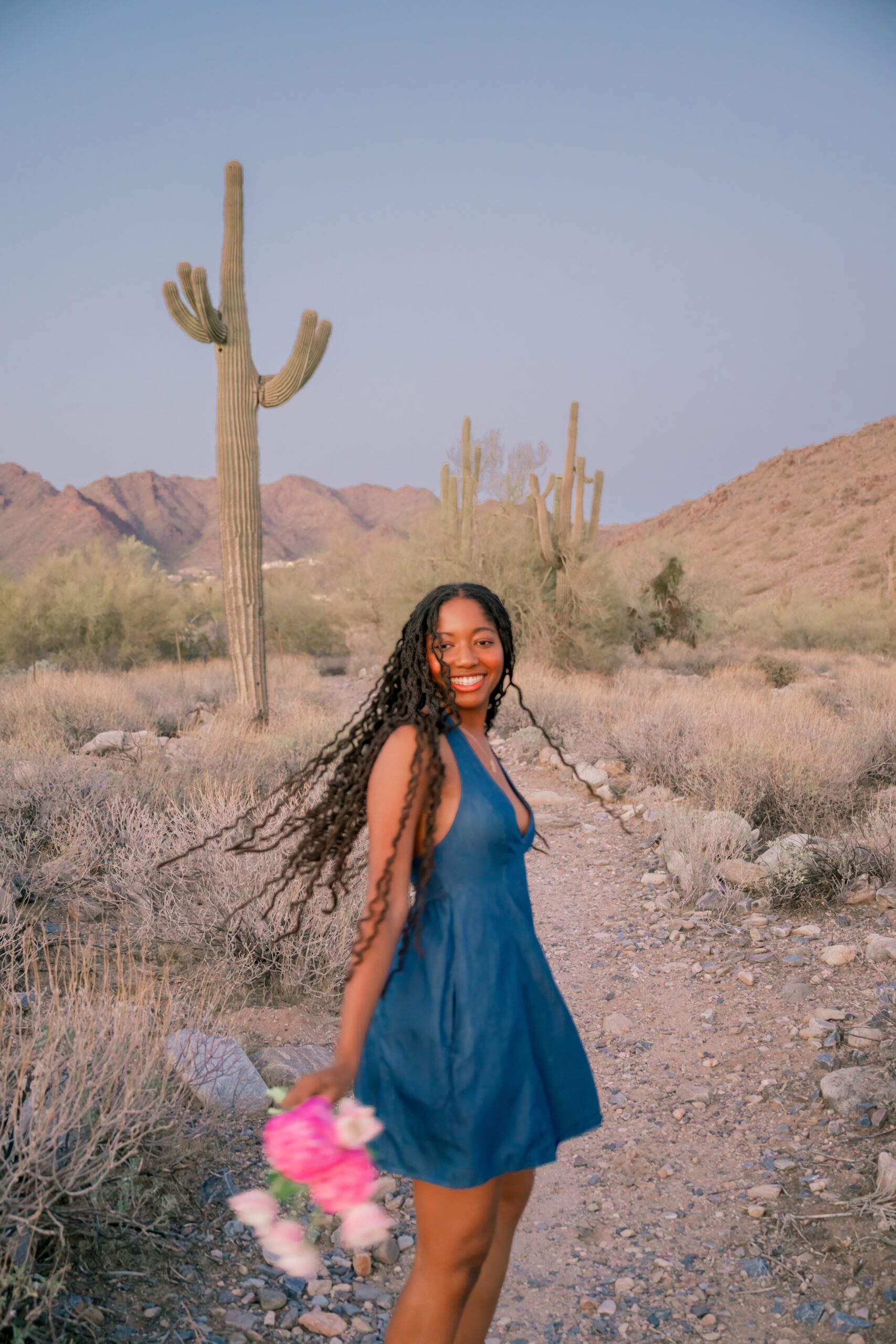 A happy woman dances in the desert in a blue dress holding pink flowers after visiting womens boutiques in scottsdale