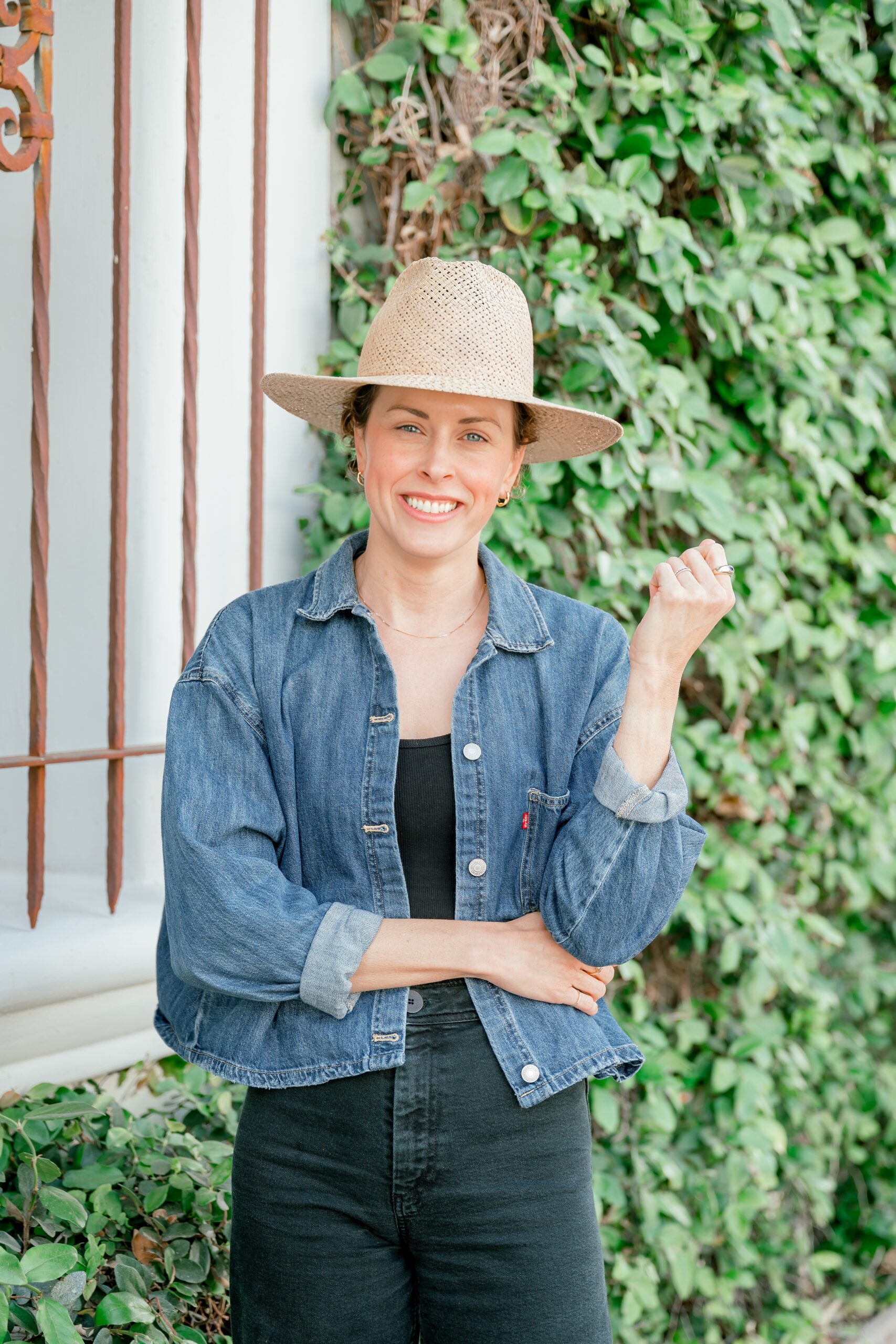 A woman stands in a denim jacket, dark pants, and woven hat by an ivy covered wall after visiting womens boutiques in scottsdale