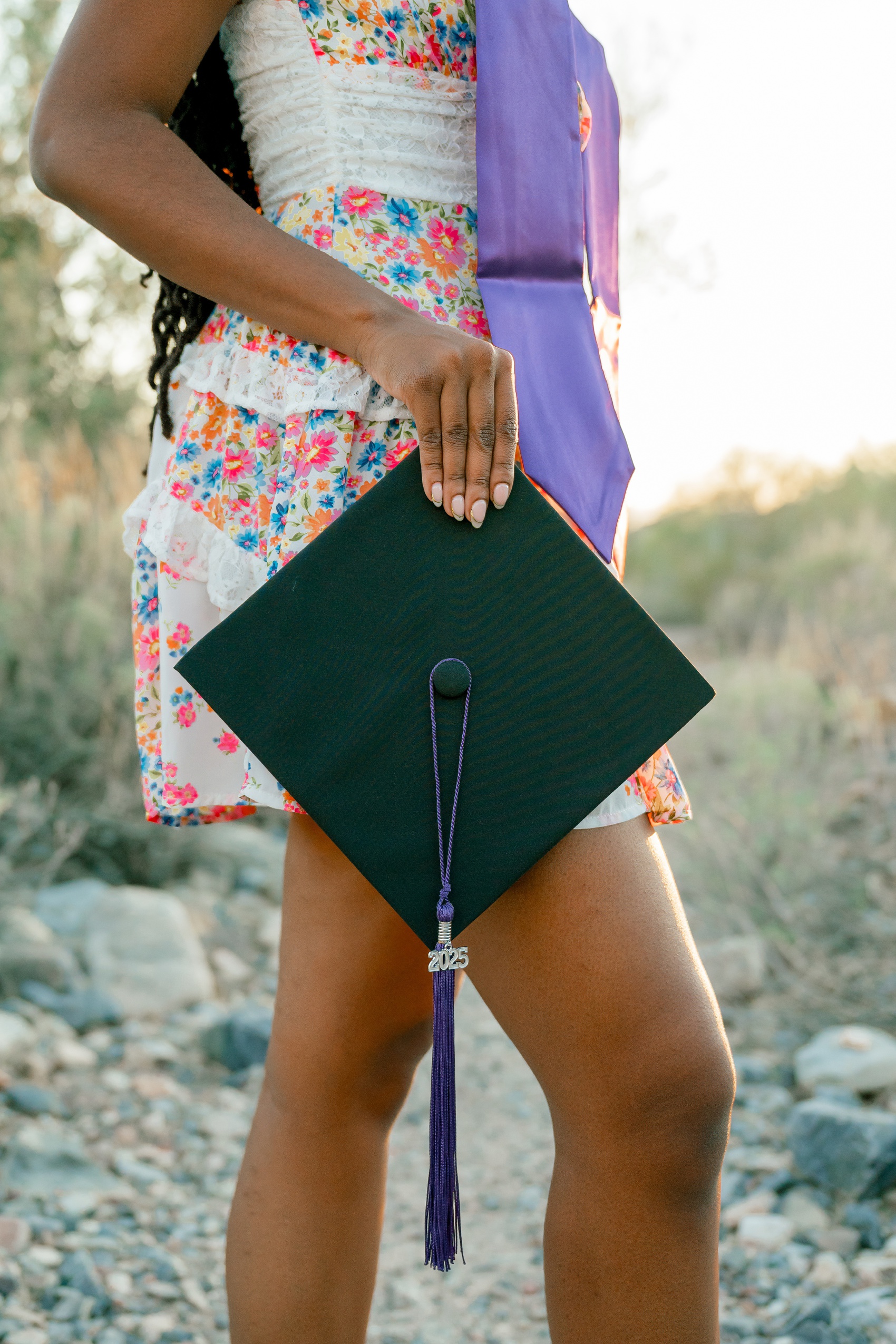 A graduate holds her 2025 graduation cap at her side in a colorful flower print dress