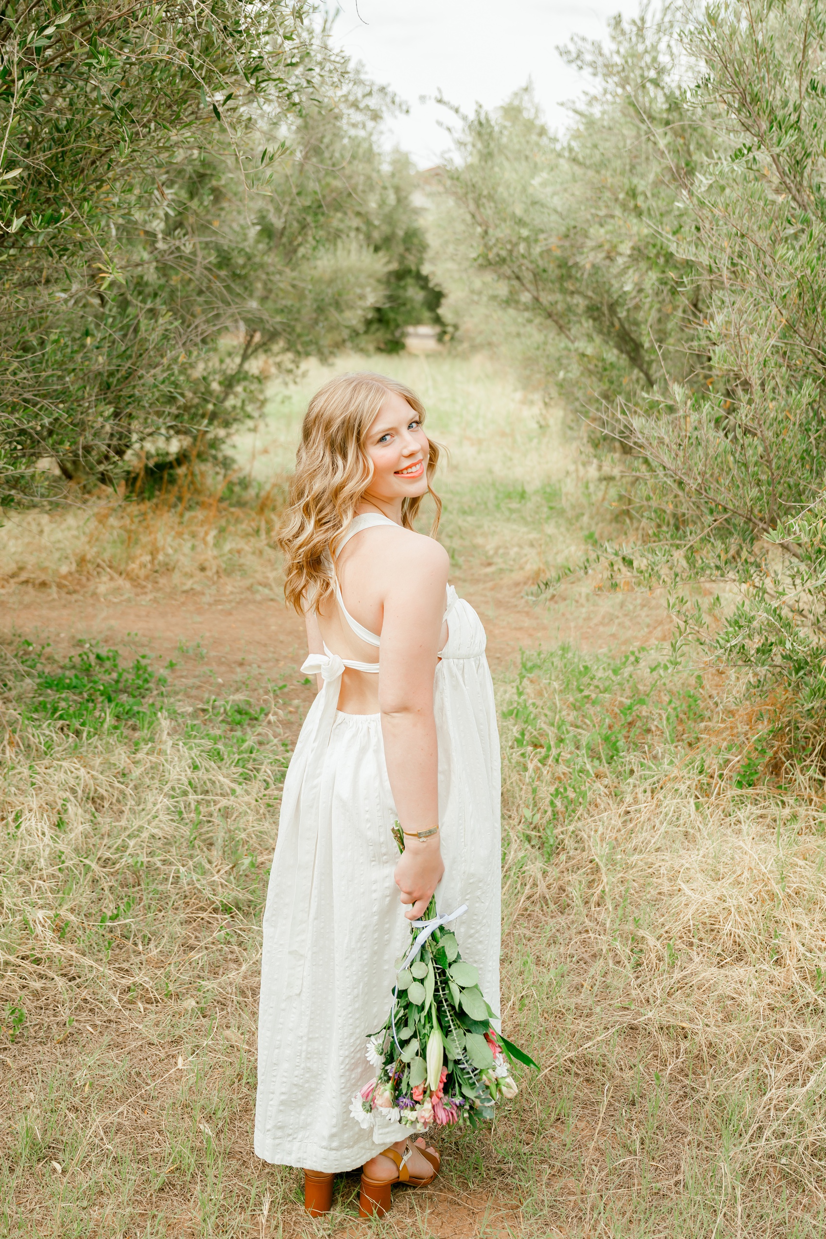 A chaparral high school senior smiles over her shoulder while walking through greenery in a white gown