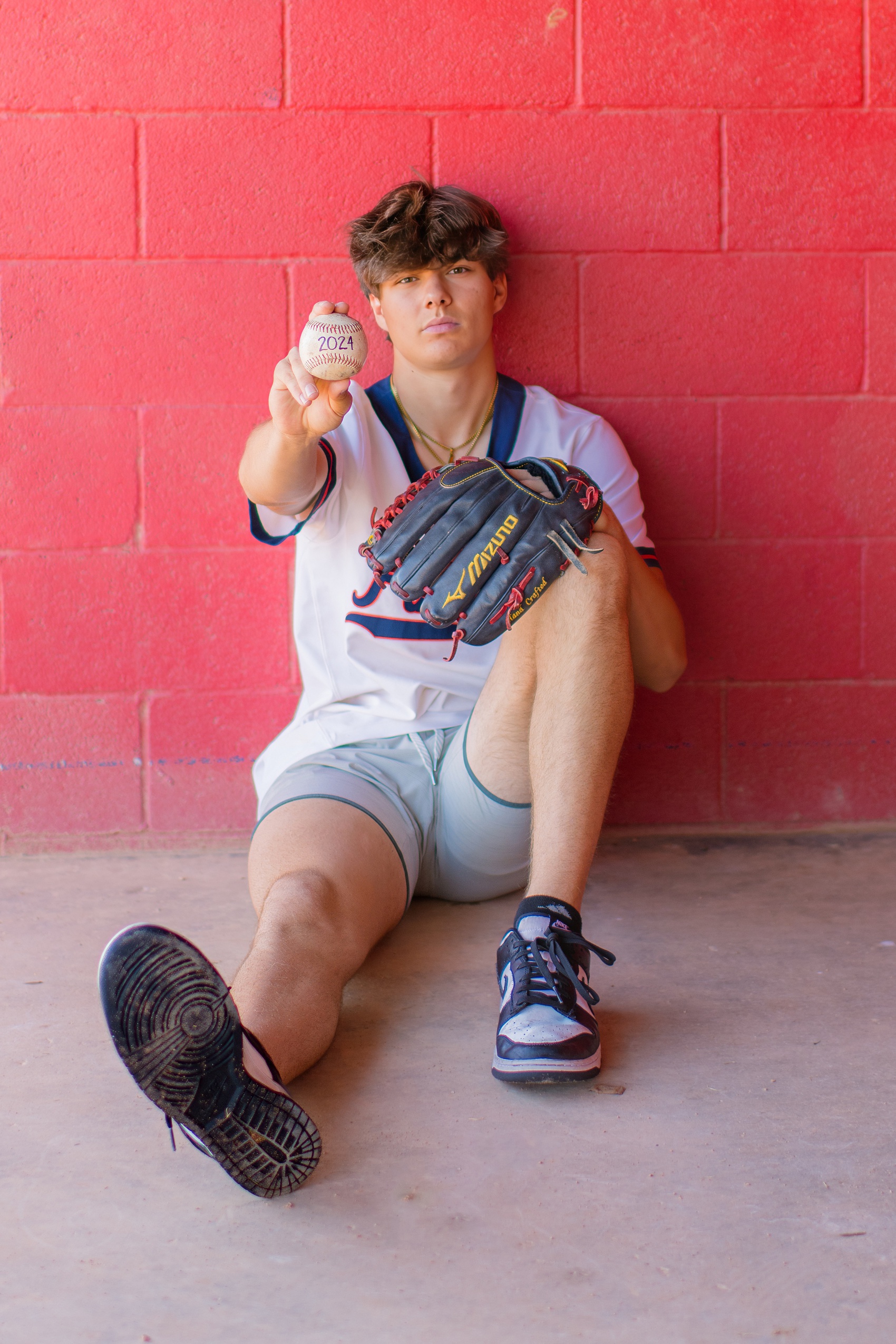 A chaparral high school senior sits against a red cinderblock wall with a baseball glove holding a ball out