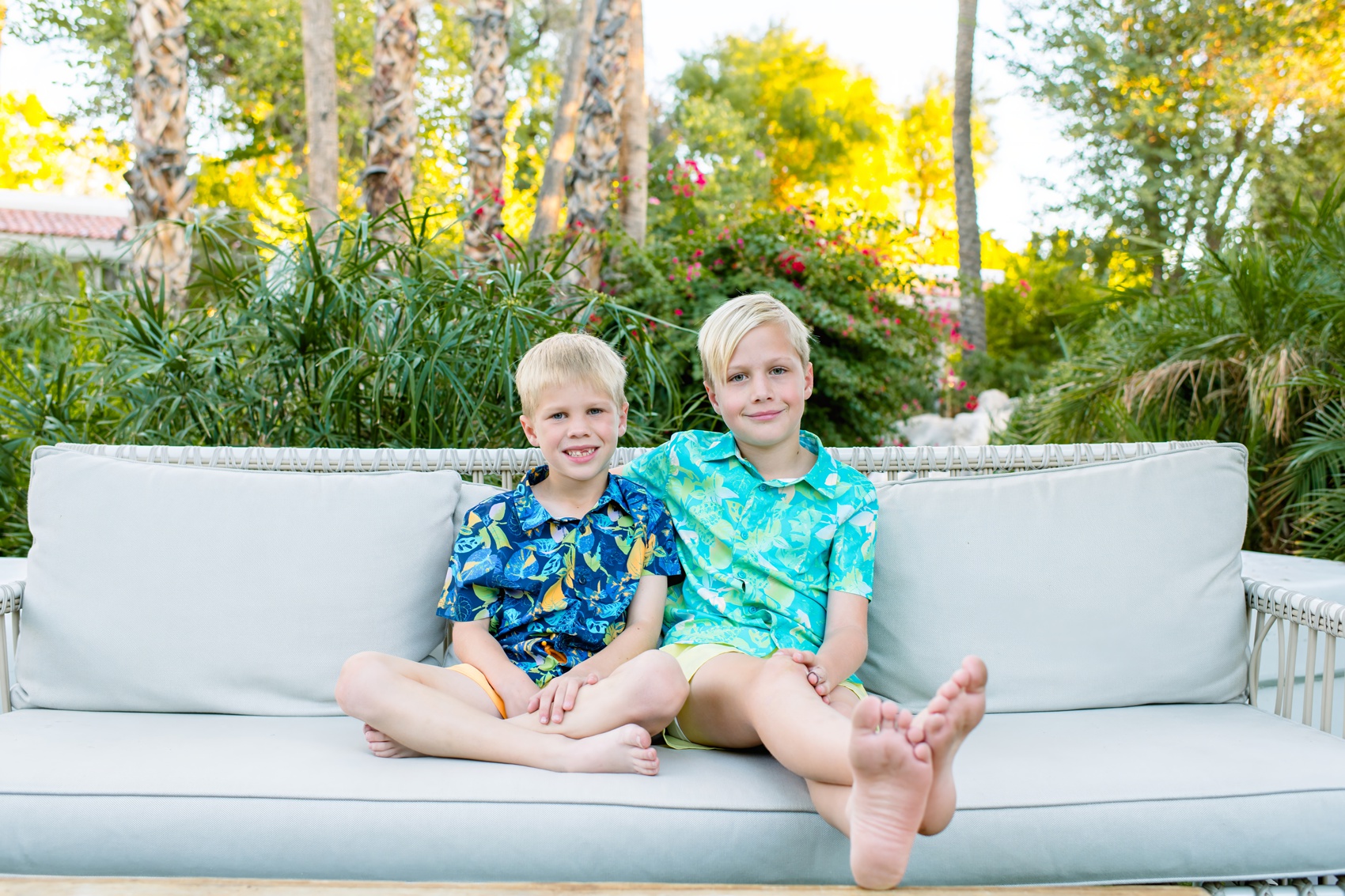 Happy young brothers smile together on a patio couch in bright tropical shirts in a garden