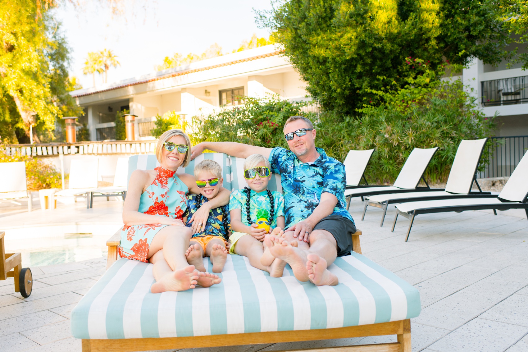A mom and dad sit on a poolside patio bed with their two young sons in matching hawaiian shirts and big sunglasses before visiting kid friendly restaurants in scottsdale